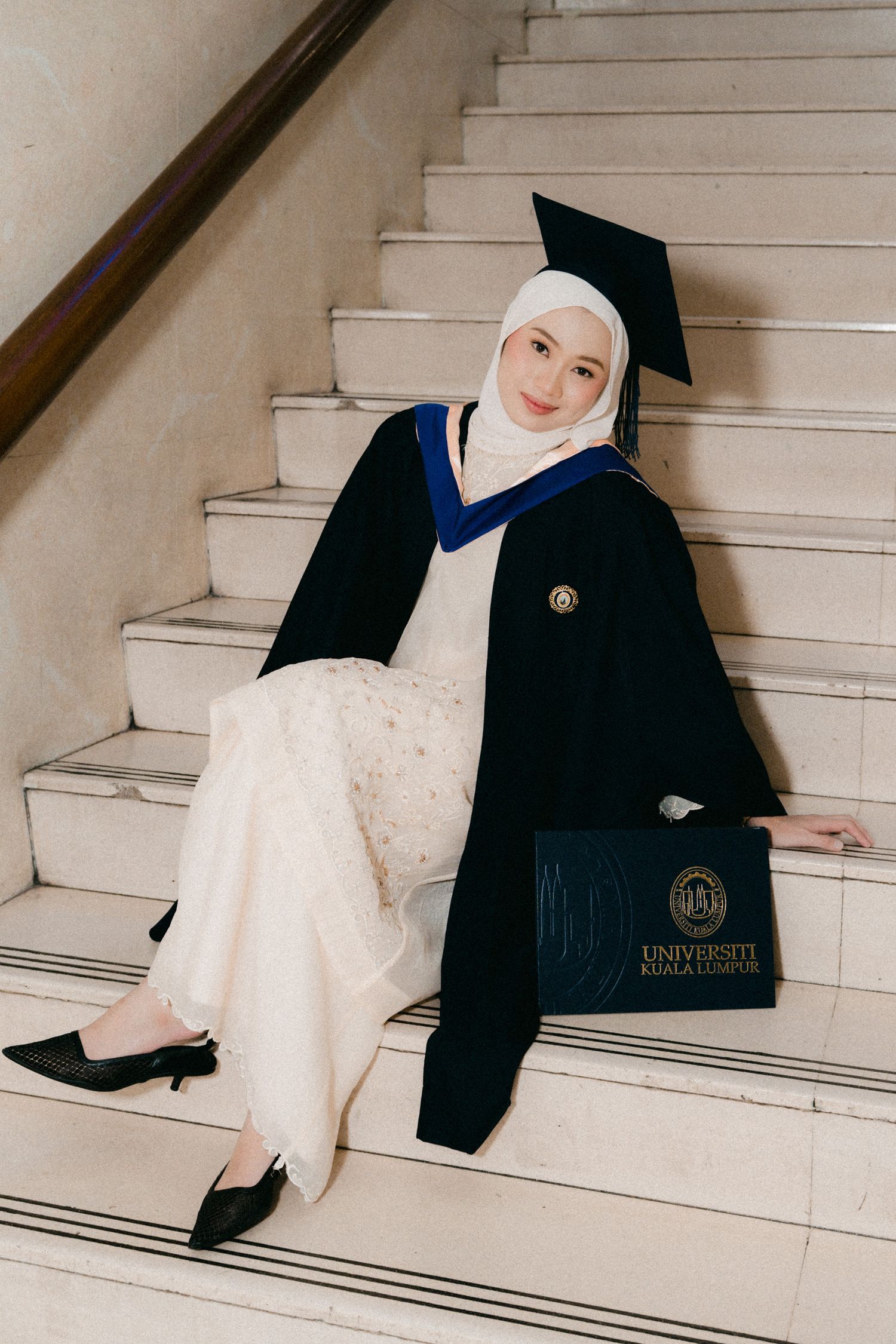 A graduation photoshoot on white stairs featuring a graduate in traditional cap and gown with white dress and hijab.