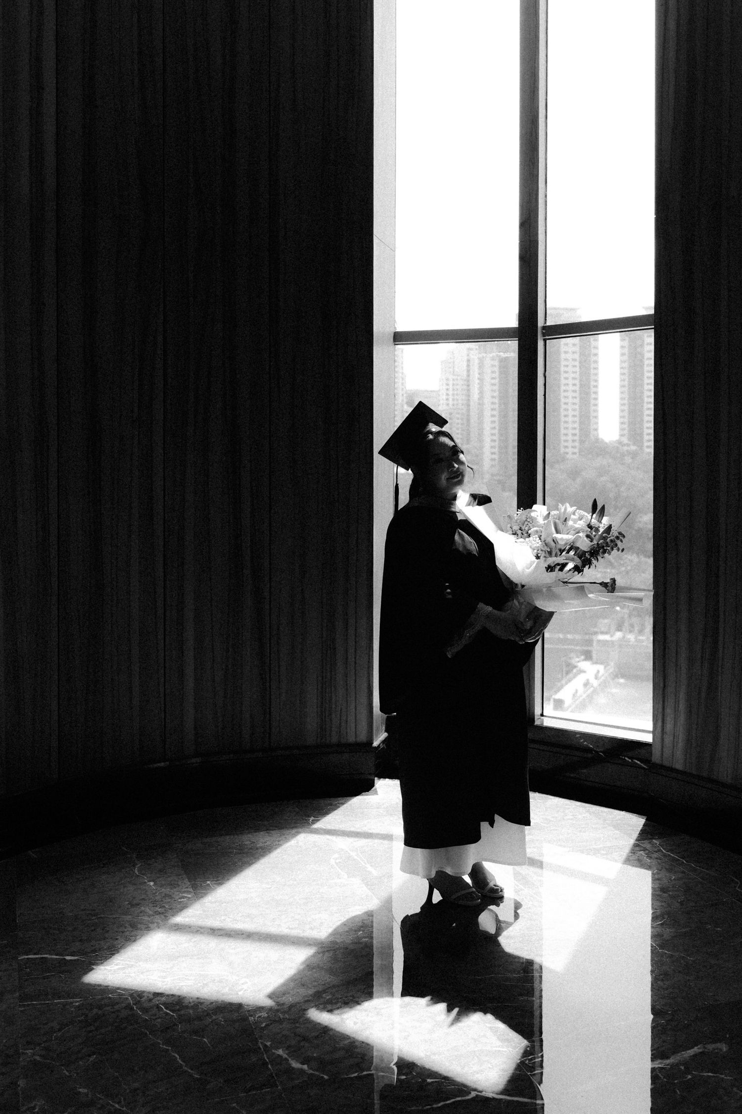 A silhouetted graduate stands in dramatic lighting by a window holding flowers in a contemplative pose.