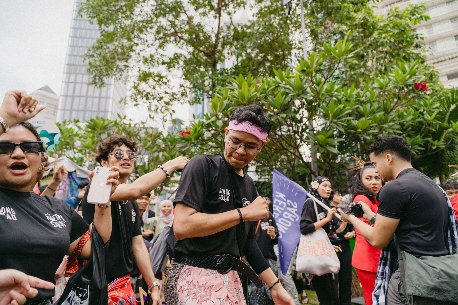 Energetic crowd gathering outdoors with tall buildings and trees in background.