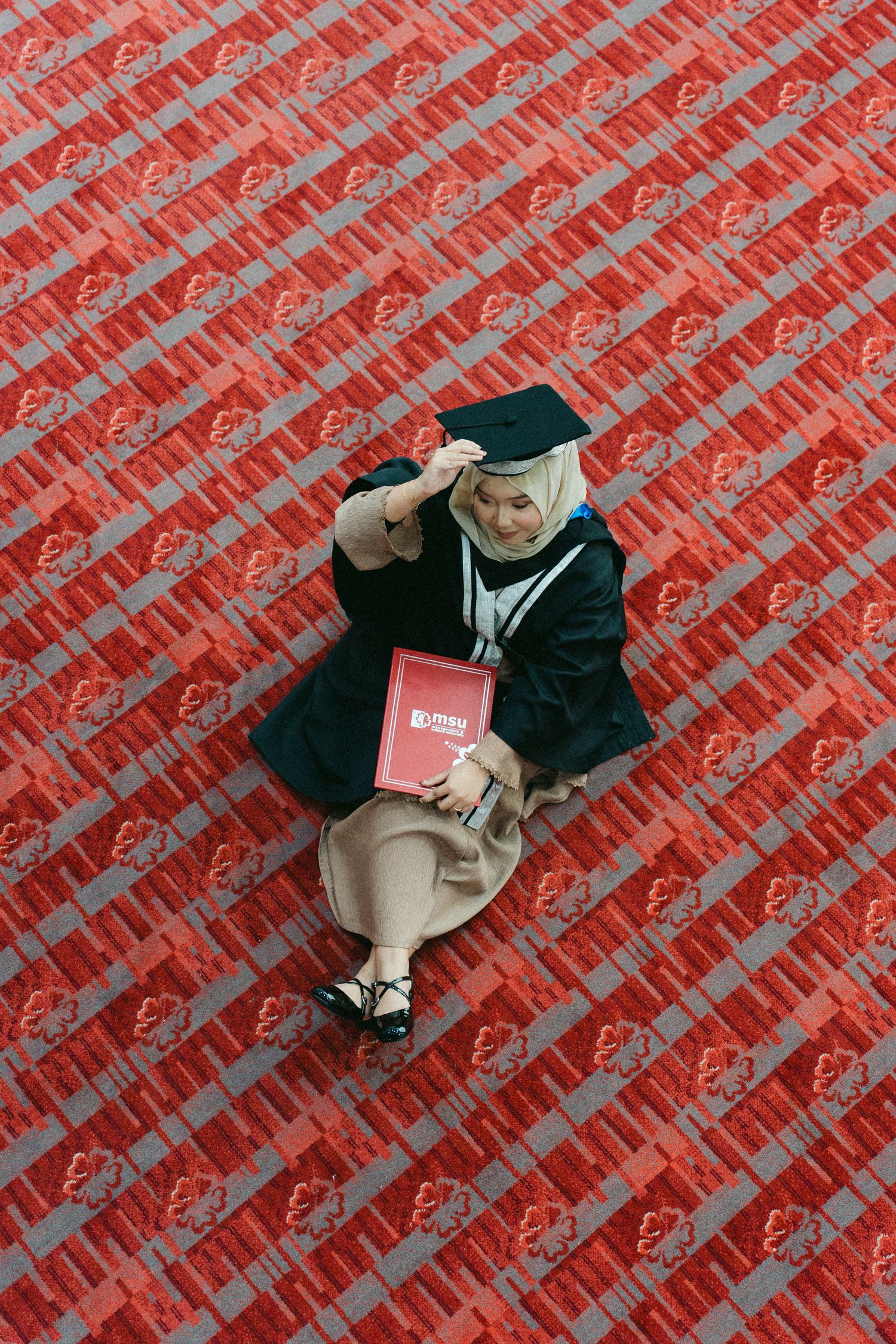 Graduate in black cap and gown sits on red textured surface holding diploma while gesturing with hand.