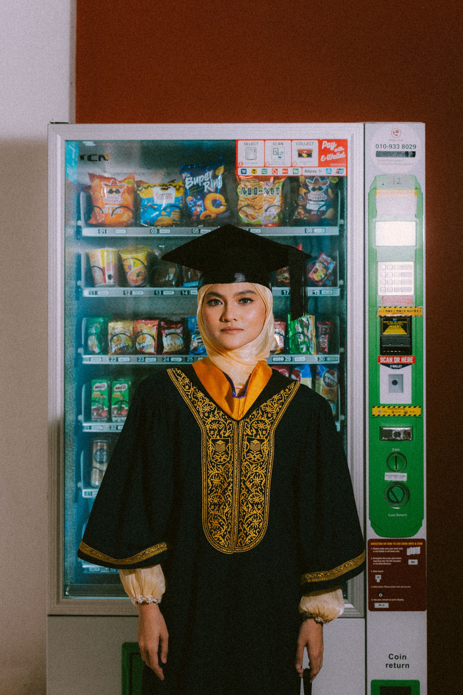 Graduate in traditional black and gold embroidered academic regalia stands in front of a vending machine.