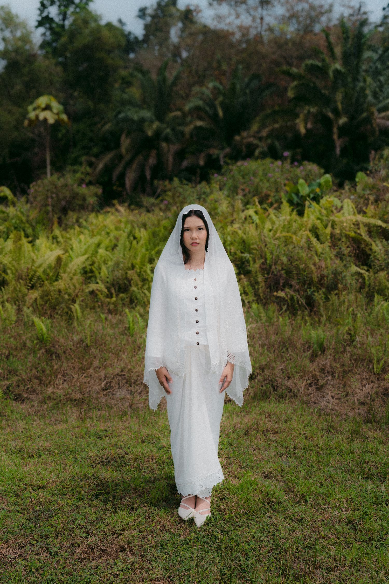 A figure in a flowing white dress and veil stands in a lush tropical garden setting.
