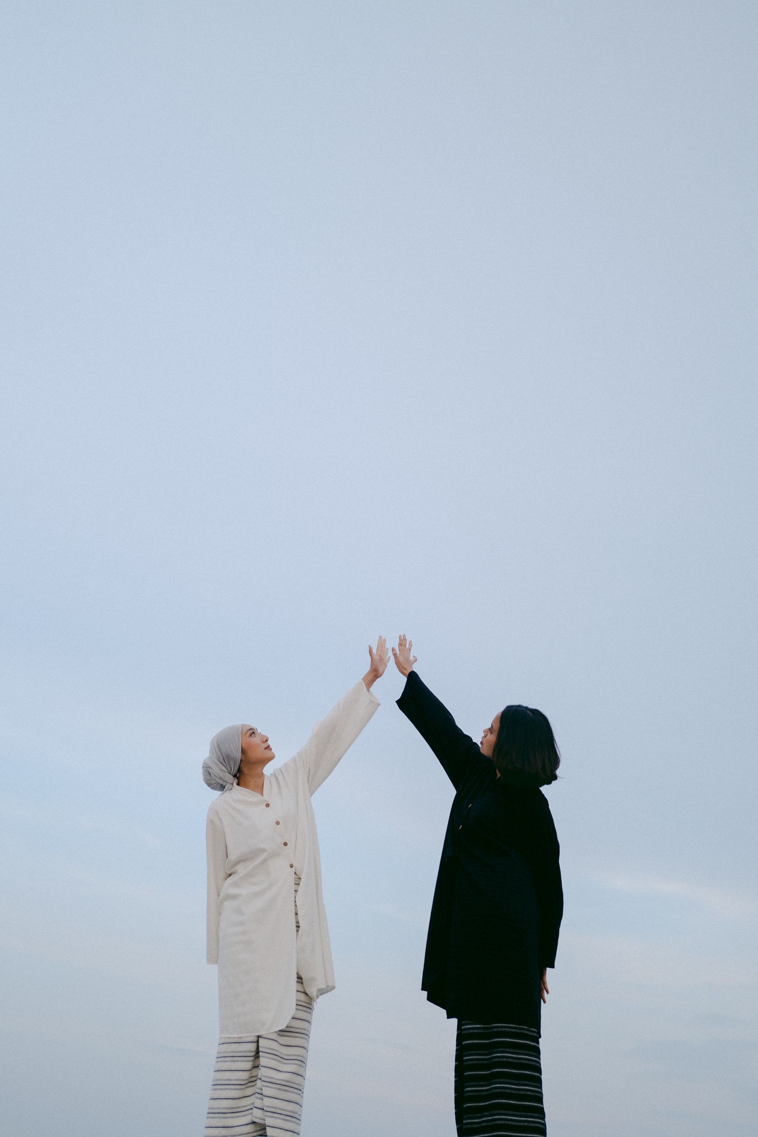 Two people in contrasting outfits reaching up towards the sky against a pale blue background.