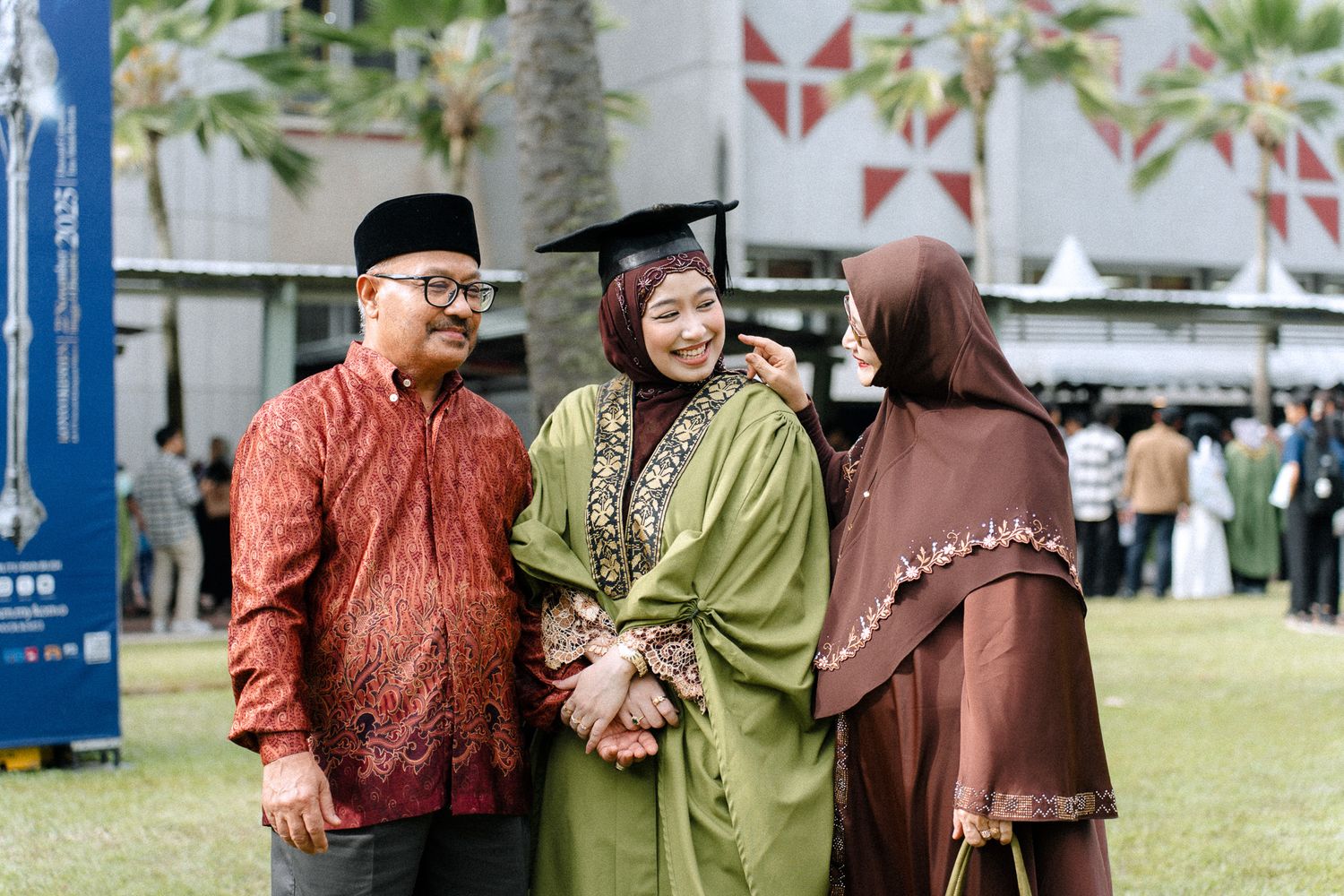 Muslim graduate in green kebaya and cap celebrating with family outdoors.