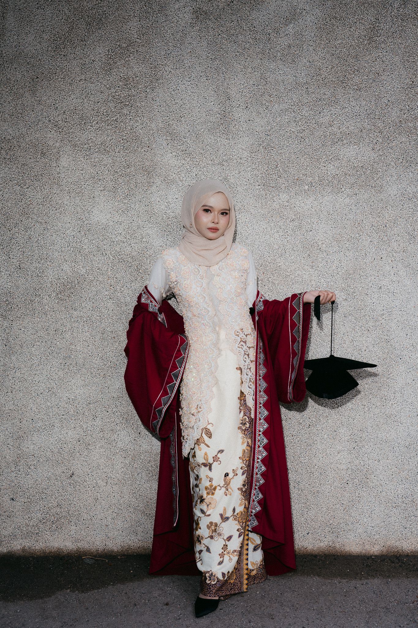 Woman in hijab wearing elegant white kebaya with floral embroidery and red robe, holding graduation cap.