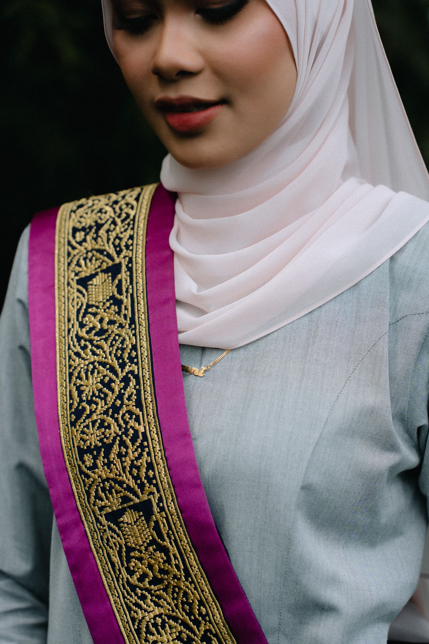 Young woman wearing a white hijab and gray outfit with an ornate purple and gold sash.