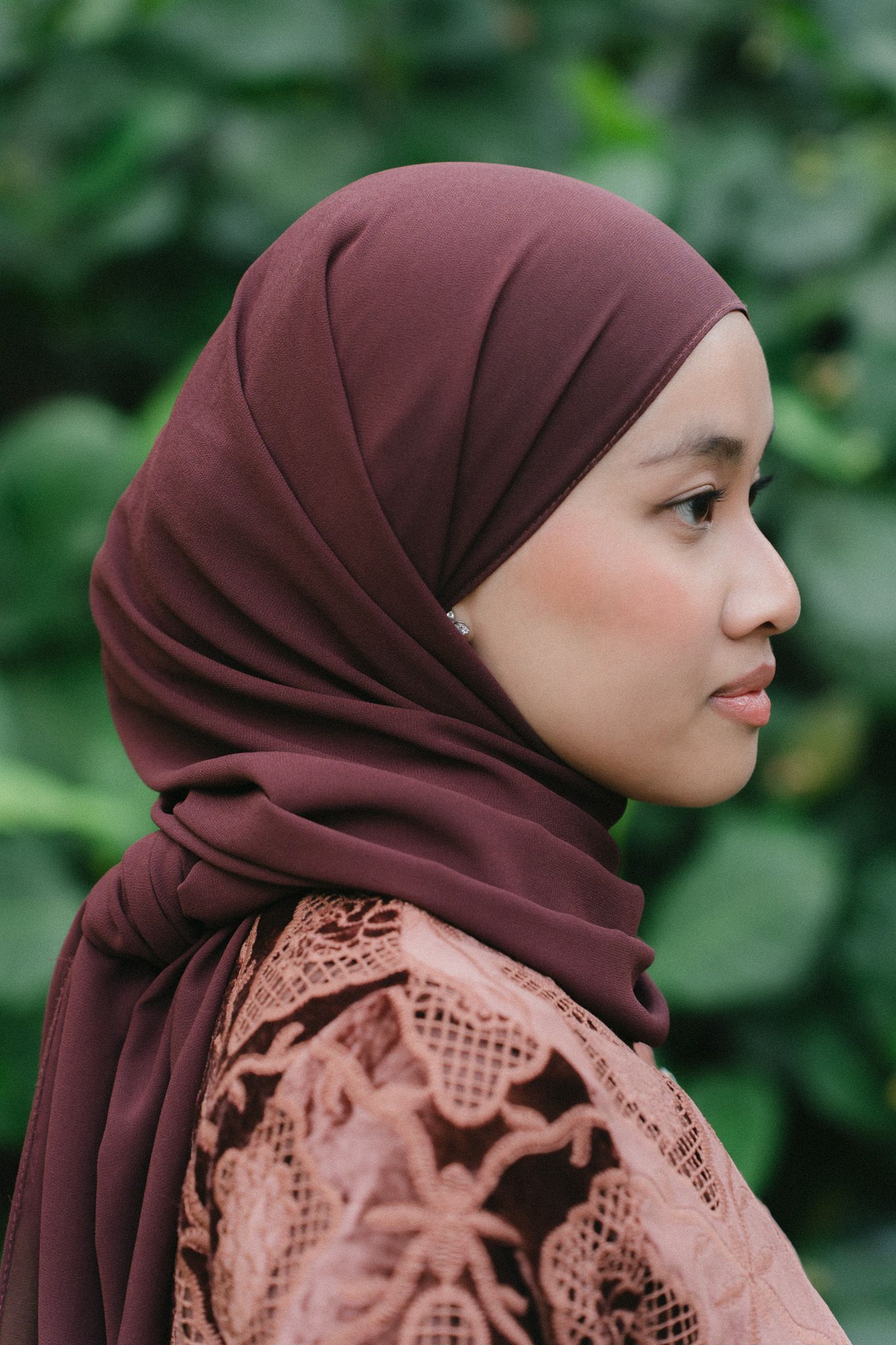Profile view of a woman wearing a dark brown hijab and pink lace top, with green foliage background.