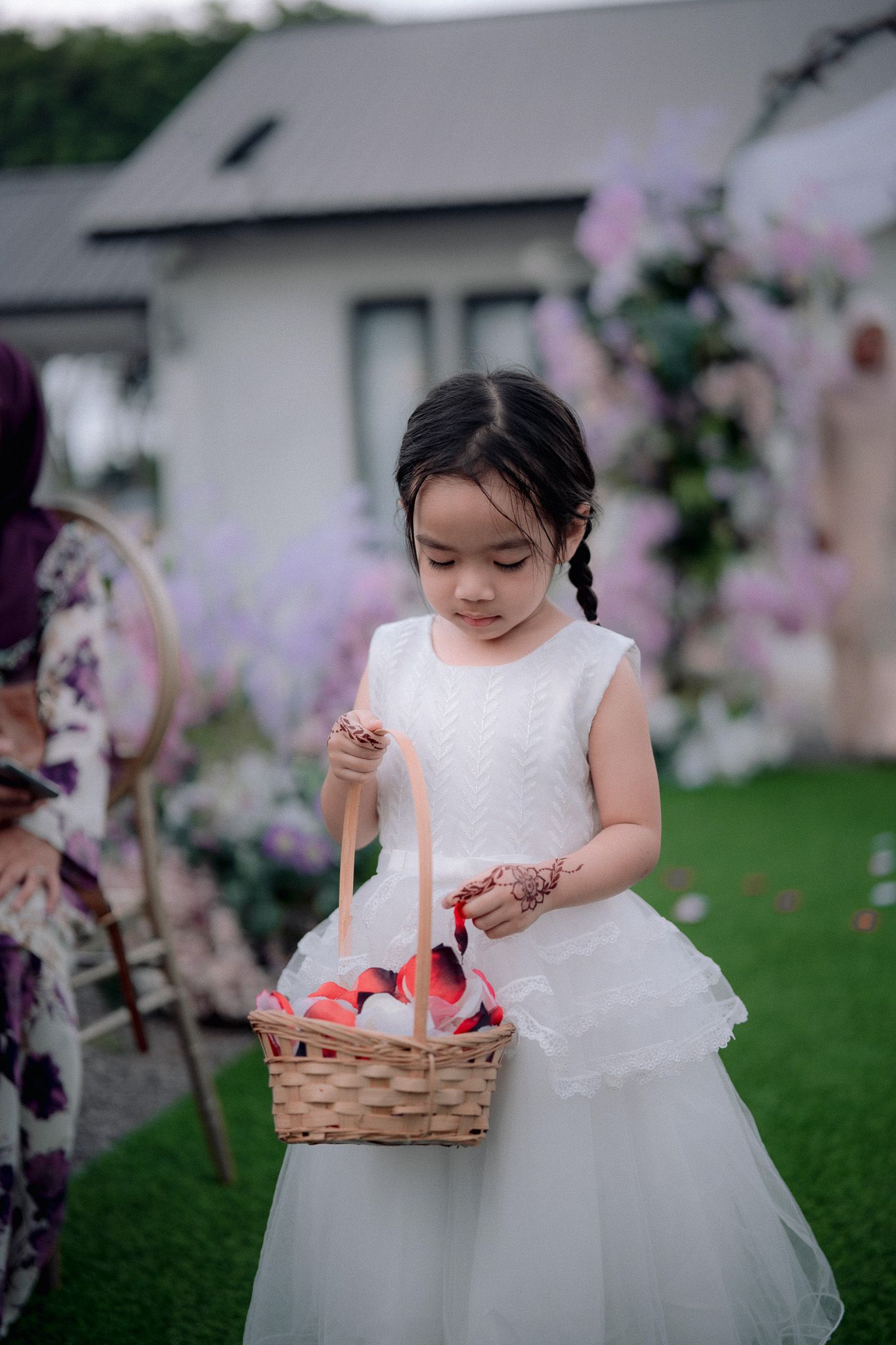 Young flower girl in white dress holding wicker basket with red petals at outdoor wedding ceremony.