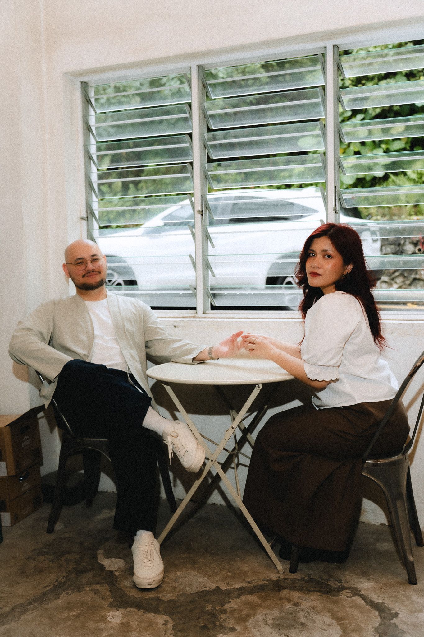A bald man and a woman with red hair sit together at a small white table near a louvered window.