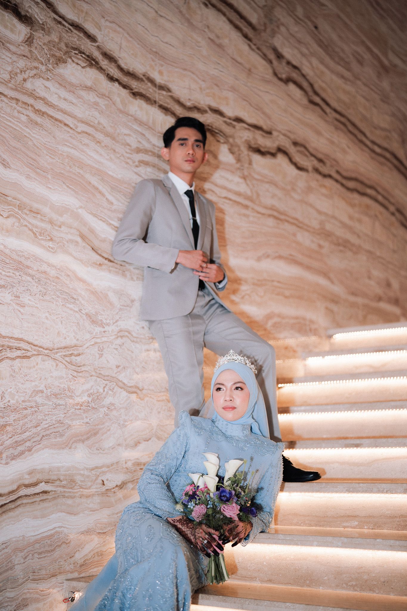 A Muslim bride in blue hijab holds flowers on stairs as groom in gray suit stands behind her.