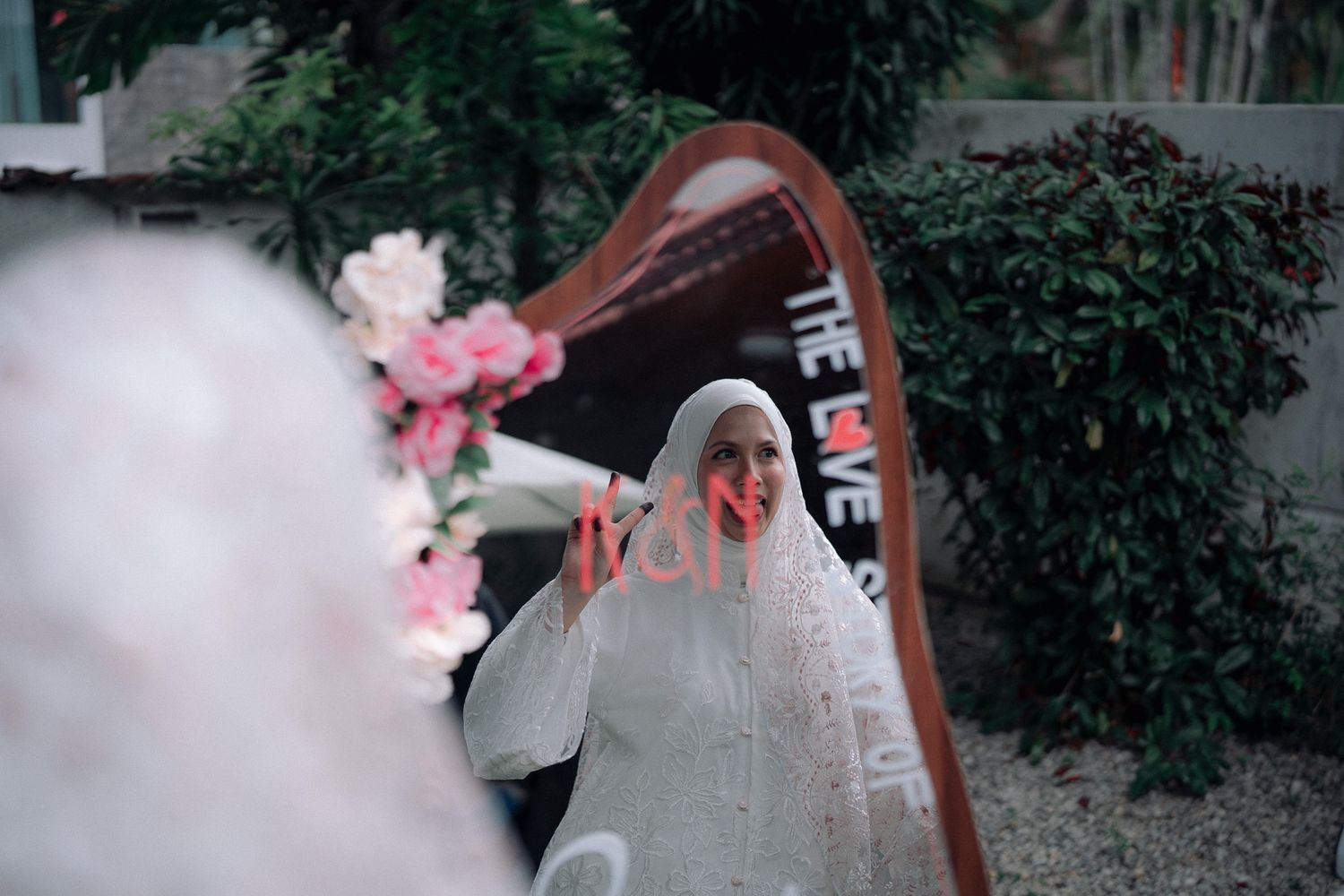 A bride in a white hijab and veil gazes into an ornate mirror surrounded by pink flowers and lush greenery.
