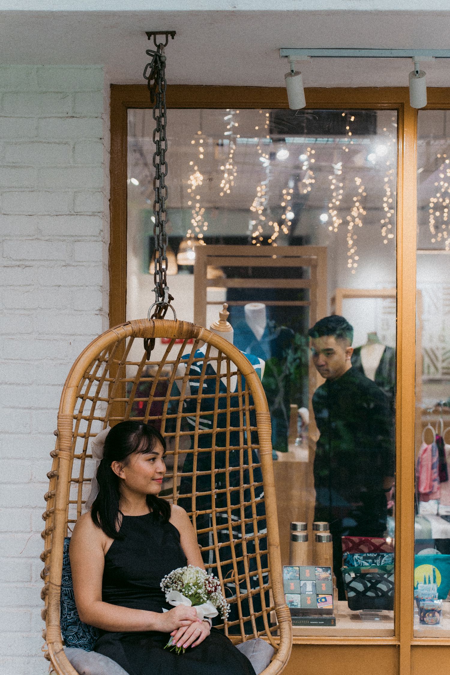 Person sitting in hanging rattan chair outside boutique storefront with string lights.