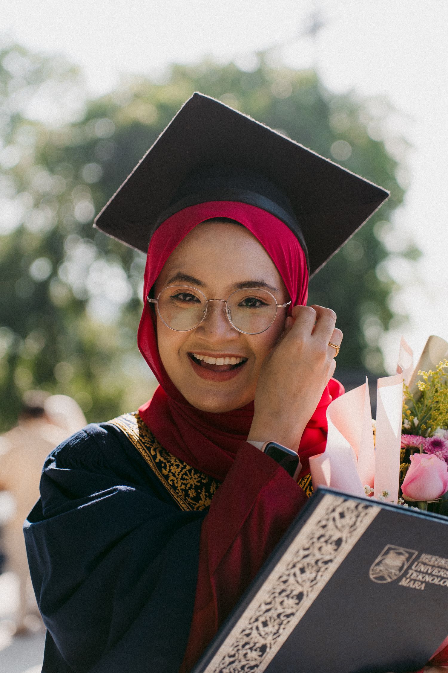 Graduate in red hijab and black graduation gown smiles while holding diploma and bouquet of flowers.