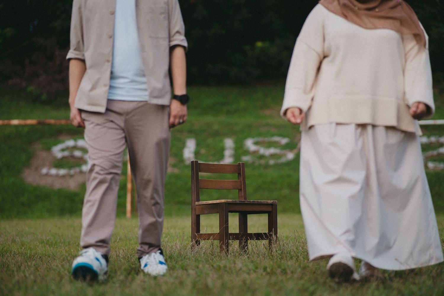 Partial view of two people in neutral colored clothing standing with wooden chair on grass.