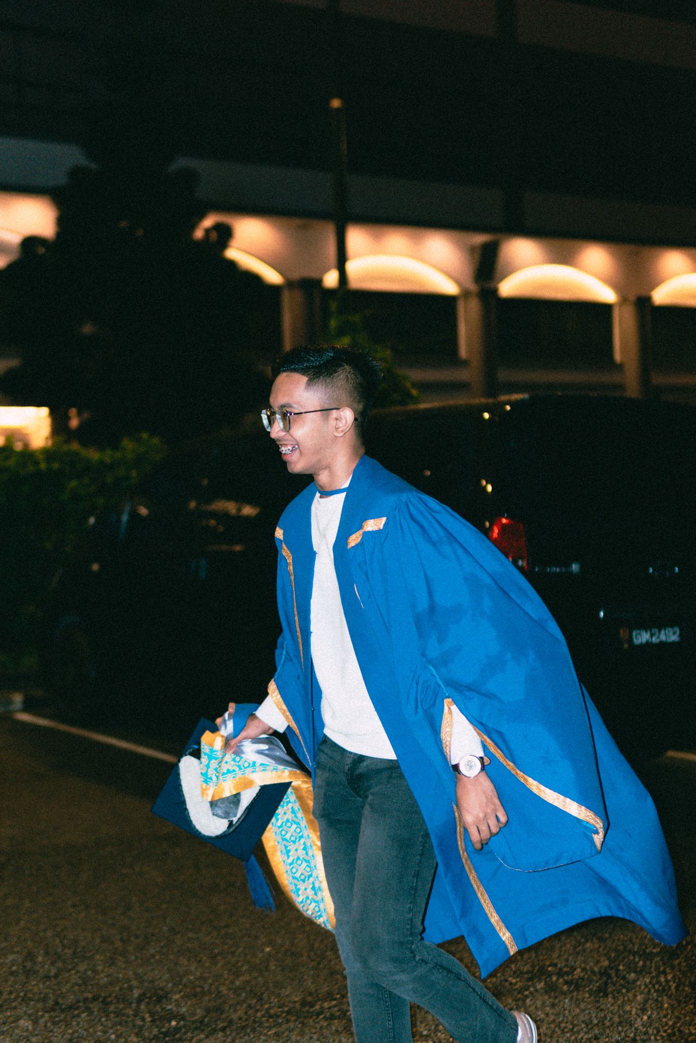 Graduate in blue gown running playfully at night in parking area.