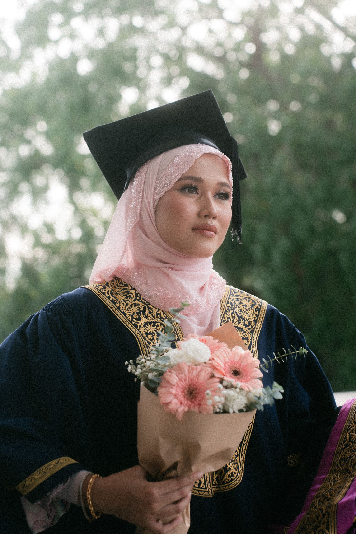 A person wearing a pink hijab and black dress holds pink and white flowers against a blurred green background.