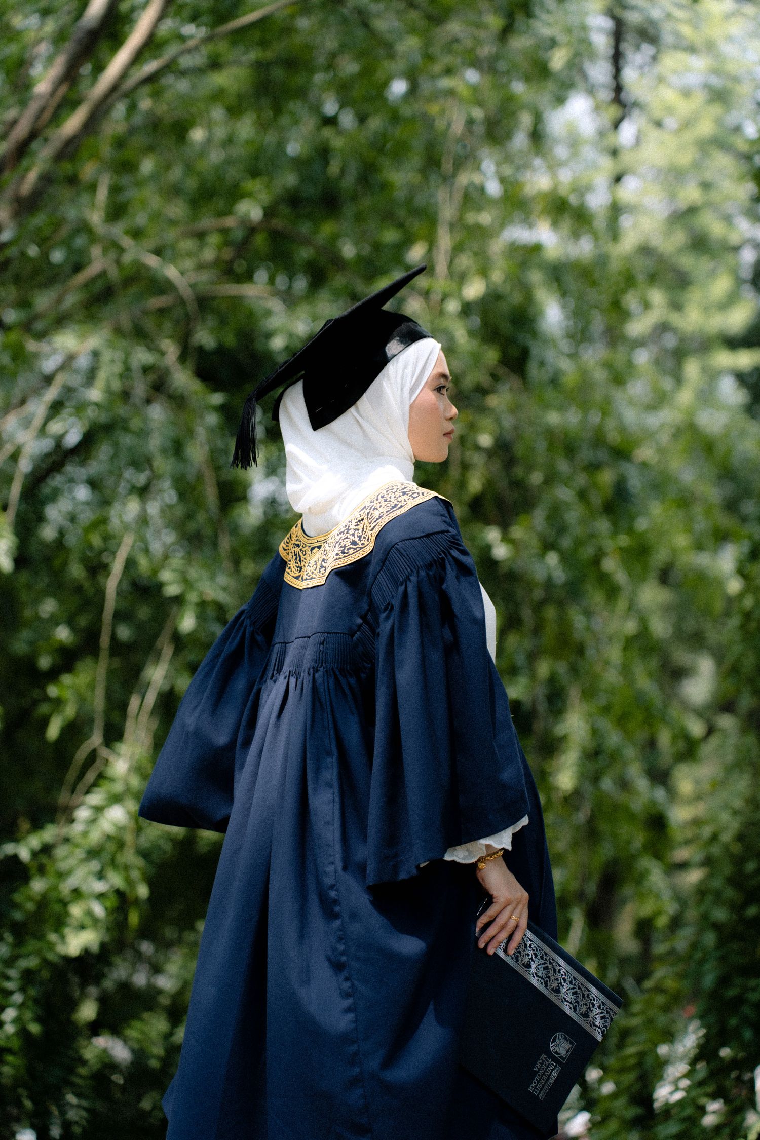 Graduate in navy blue academic gown and white hijab holds diploma while standing in front of green foliage.