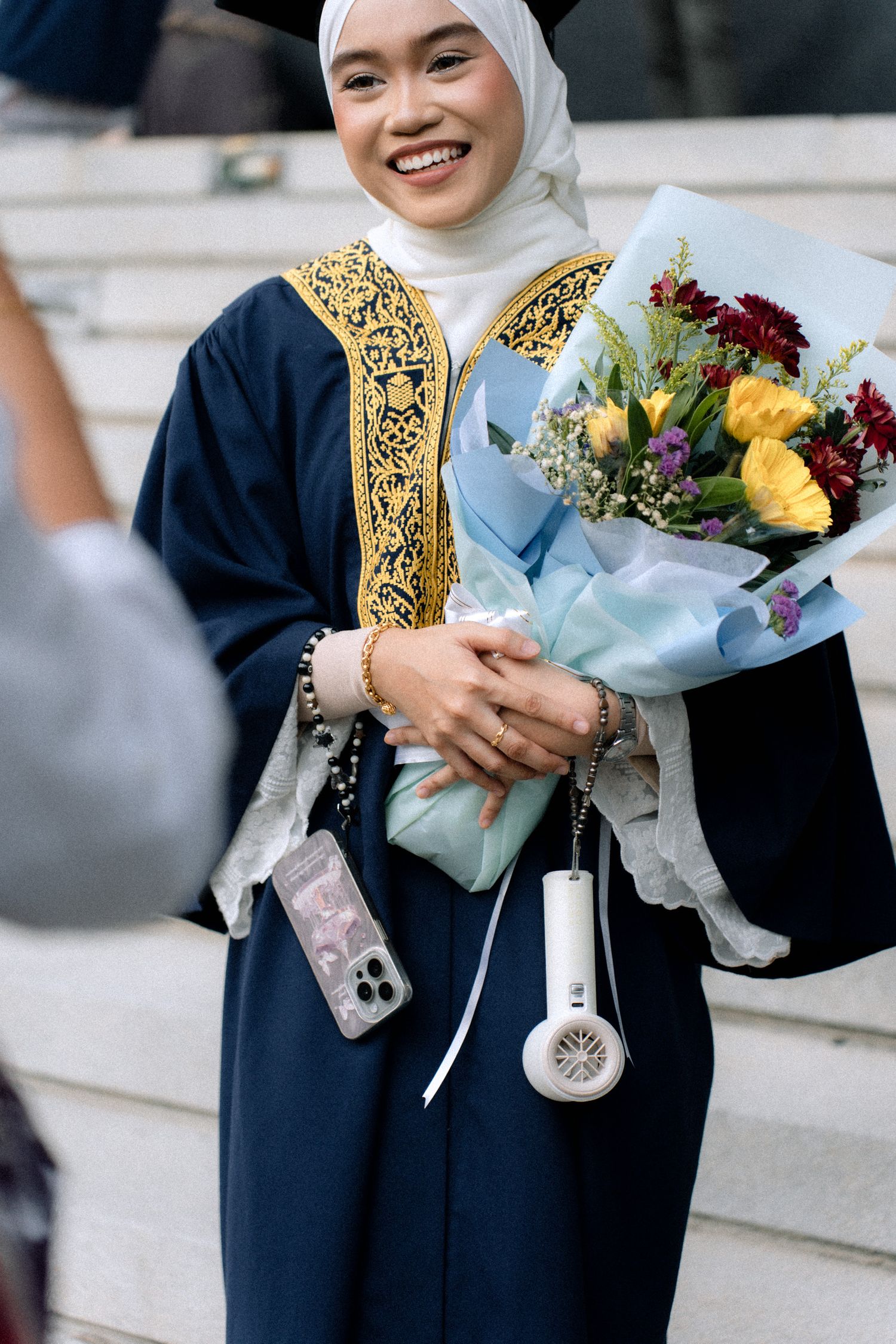 Graduate in navy blue academic regalia with gold trim holds colorful flower bouquet during commencement ceremony.