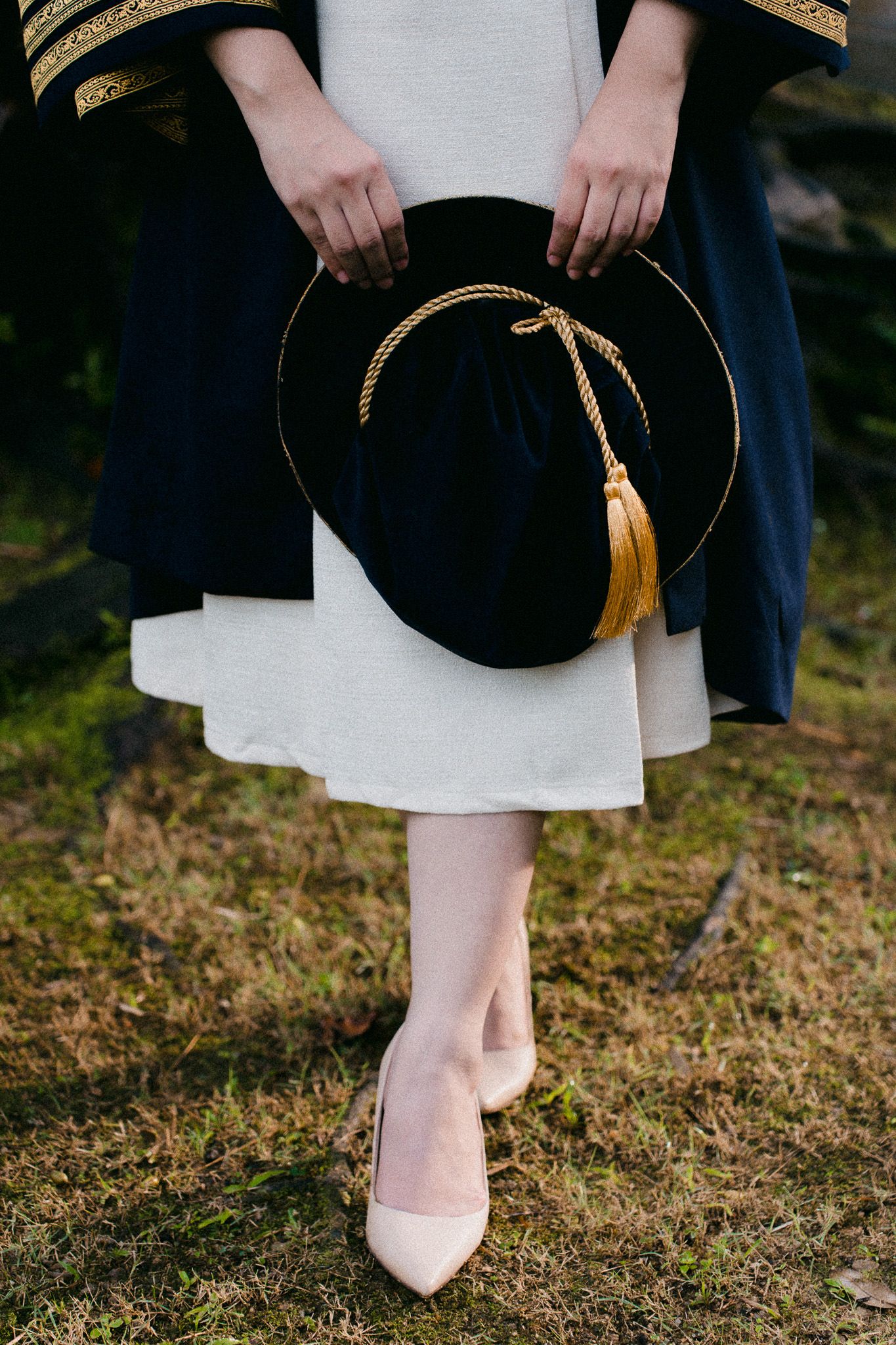 Graduate holding a black doctoral cap with gold tassel, wearing a white dress and pink heels on grass.