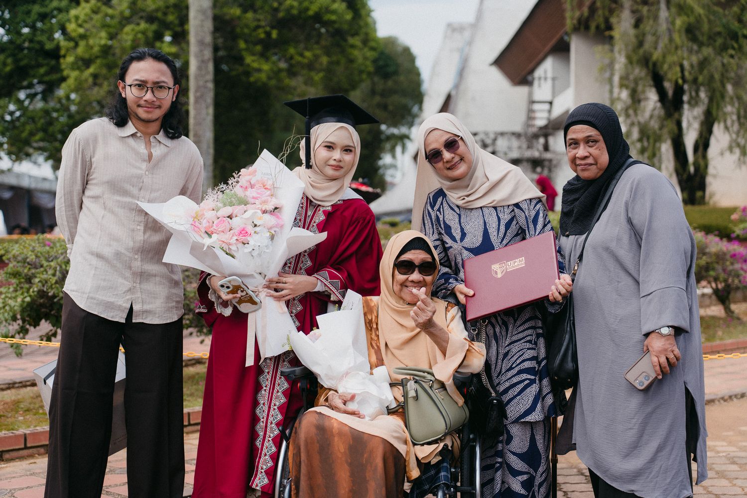 Group of Muslim women celebrating graduation outdoors, graduate in cap and gown holds flowers beside woman in wheelchair.