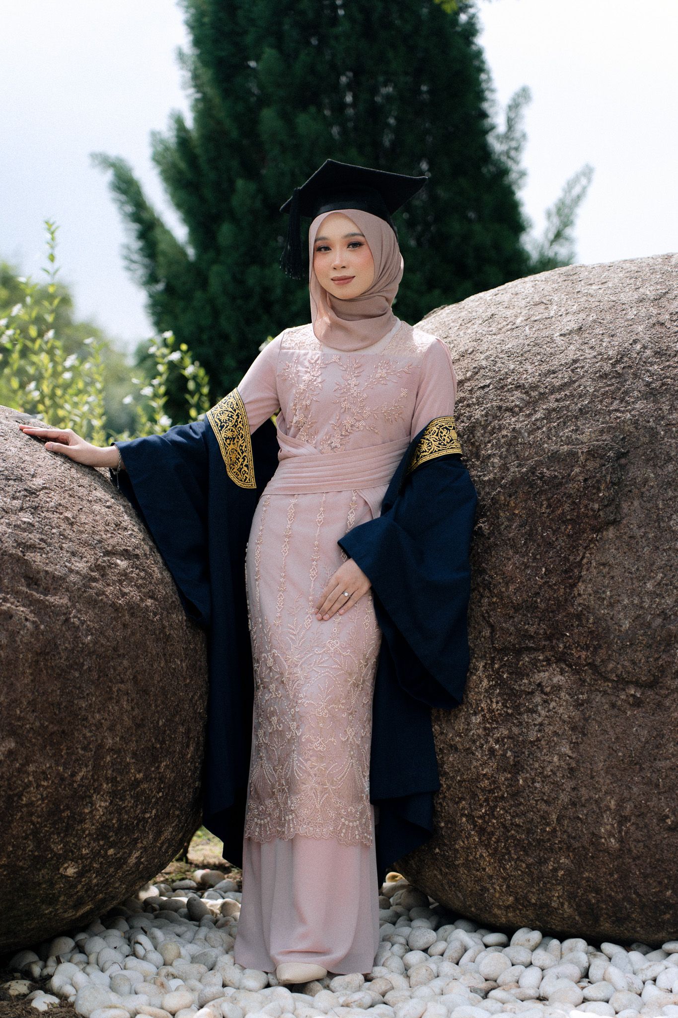 Graduate in pink hijab and dress holding cap, standing between large rocks on white pebbles with trees in background.