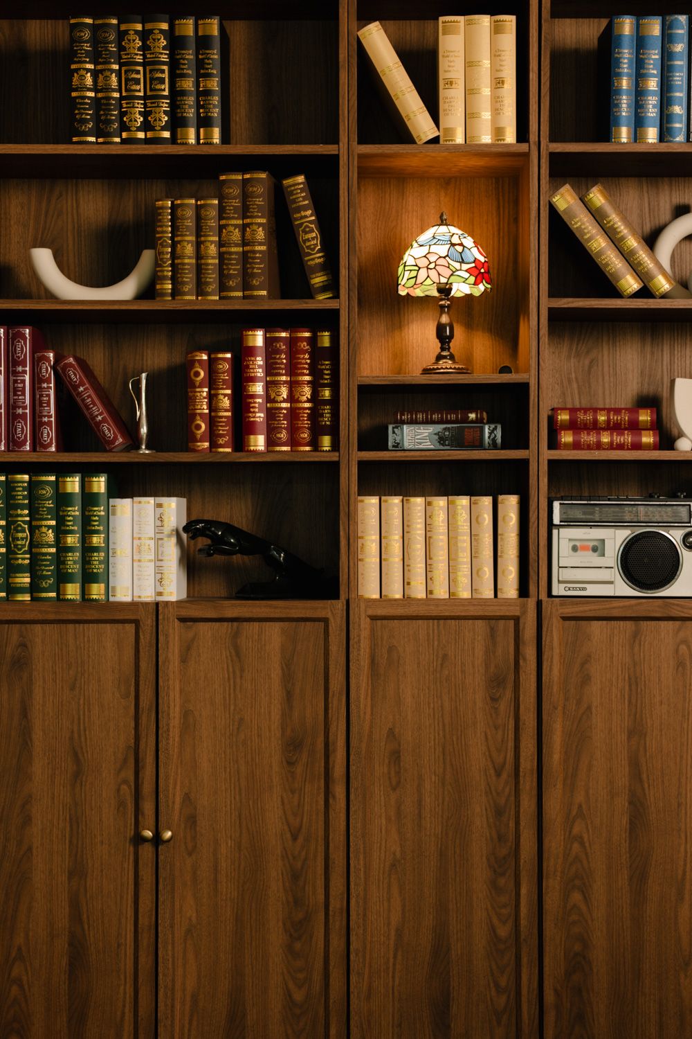 Wooden bookshelf with vintage books, Tiffany lamp, retro radio, and decorative items in a classic library setting.