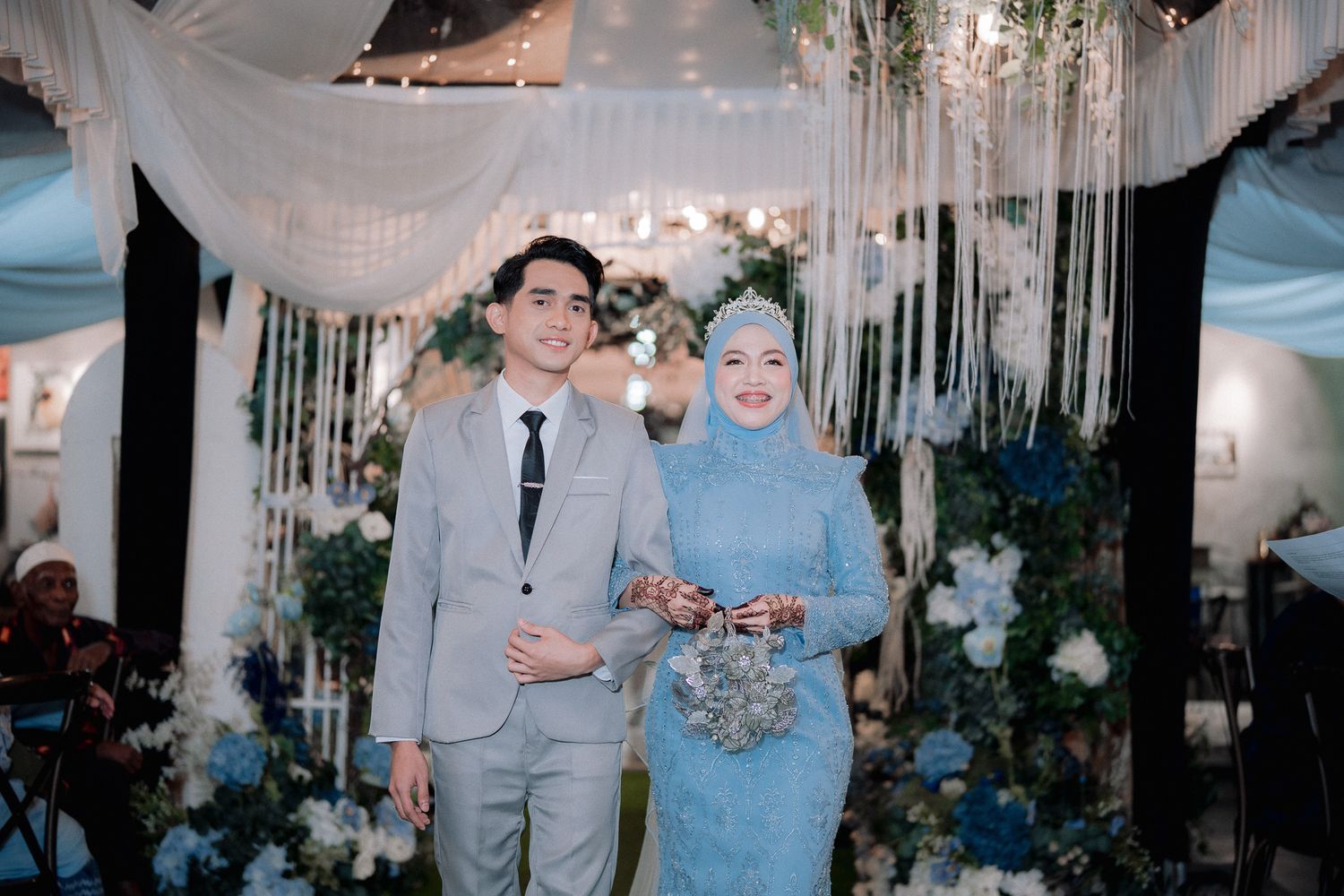 A couple in elegant blue outfits poses at a floral wedding ceremony with white draping and flower arrangements.