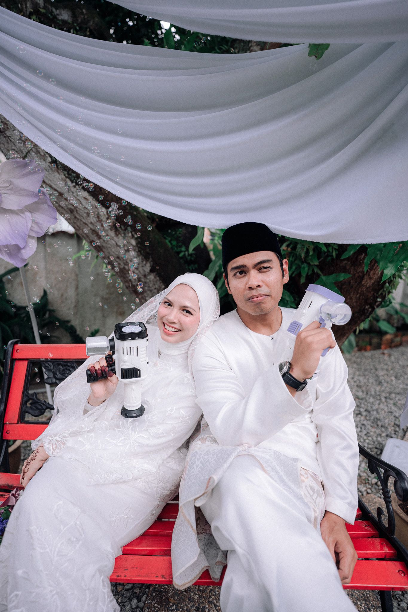 Muslim couple in white traditional attire sitting on red bench, holding tools, smiling outdoors under draped fabric.