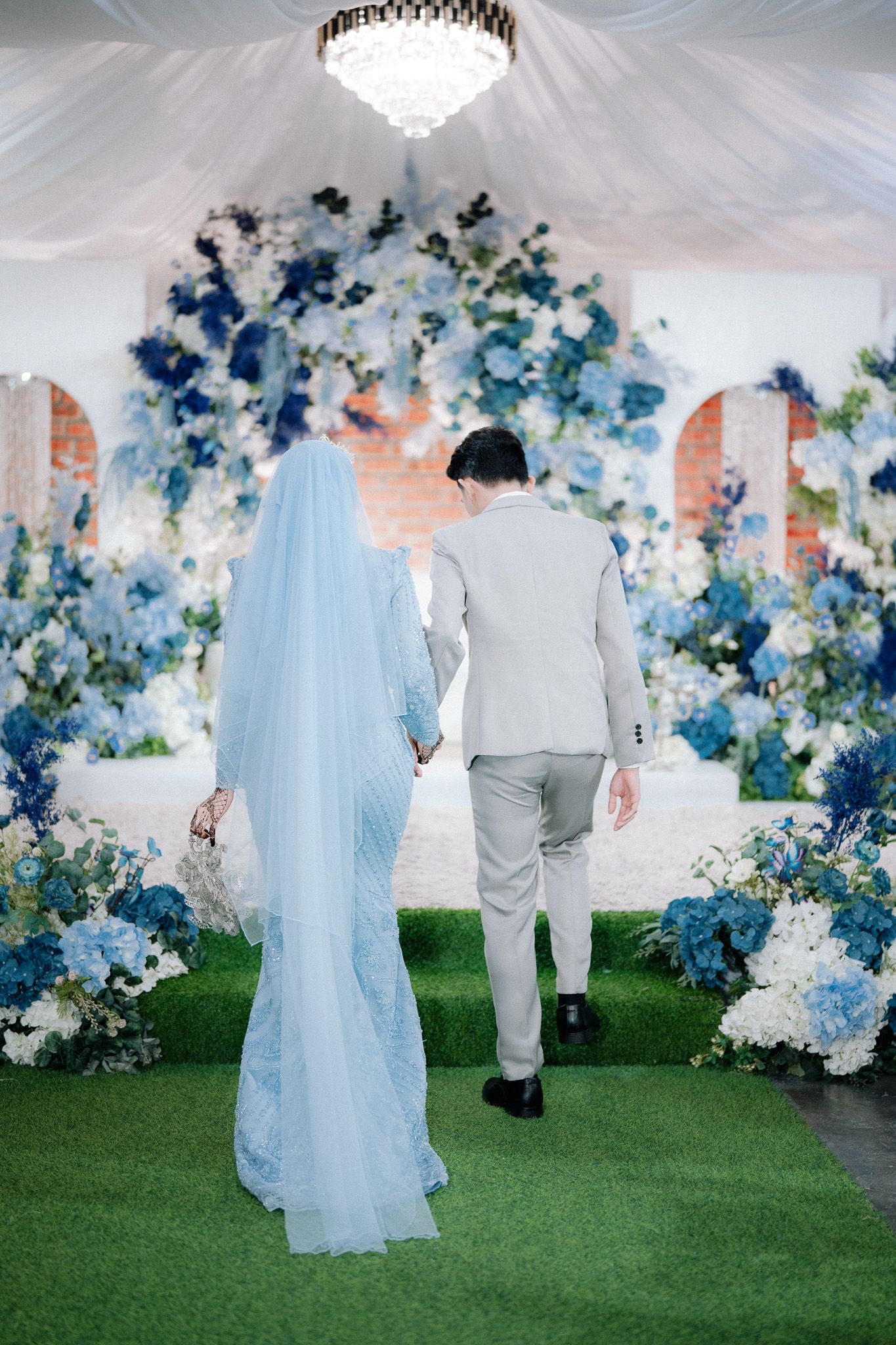 Bride and groom walking hand in hand toward stunning blue and white floral arch at romantic wedding venue.