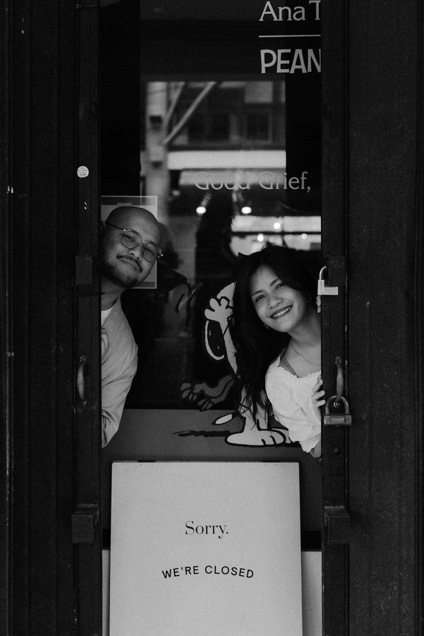 Two people peek through a closed storefront door with a Sorry We're Closed sign in black and white.