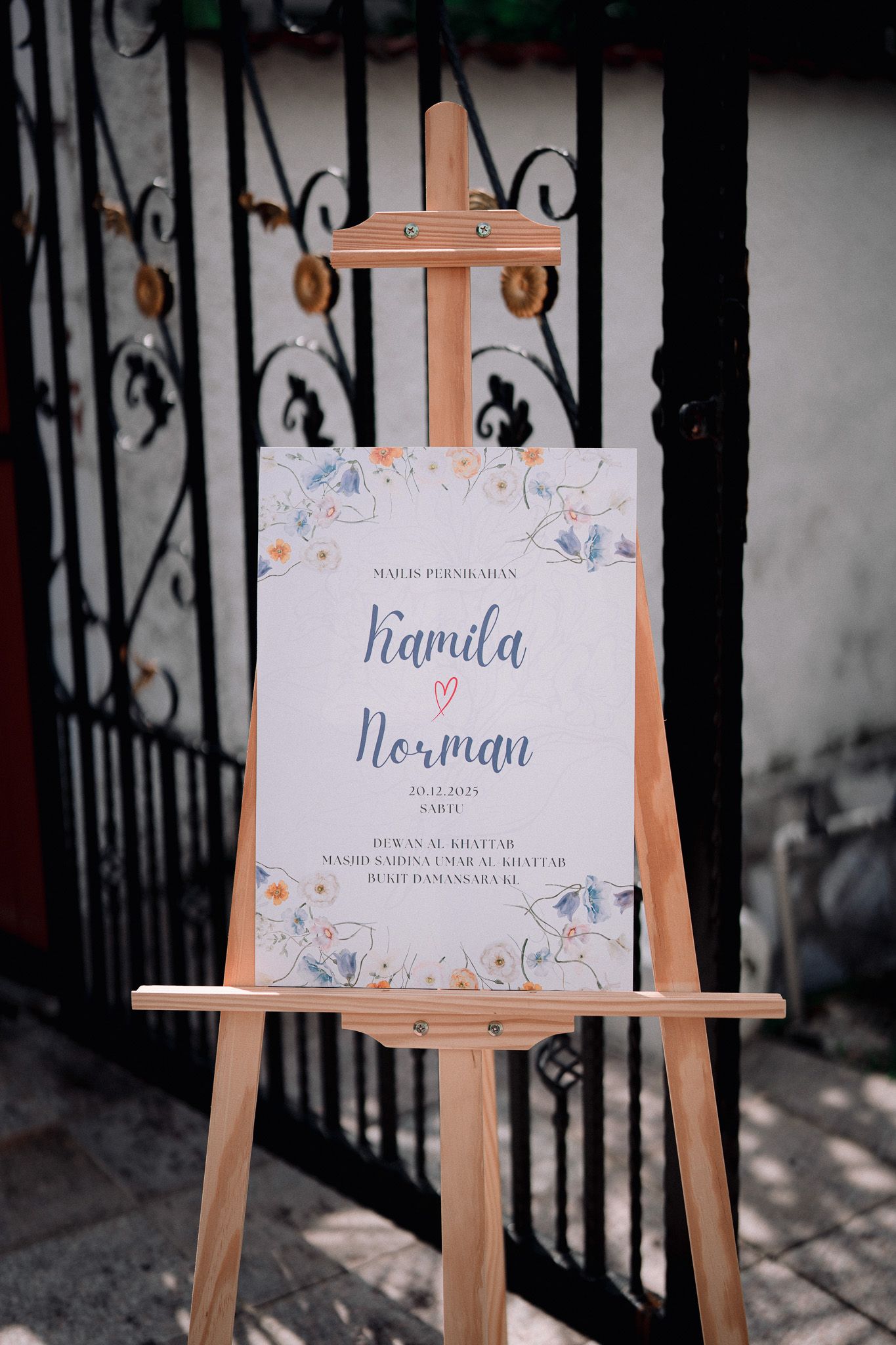 Wooden easel displaying a floral wedding welcome sign for Kamila and Norman outdoors.