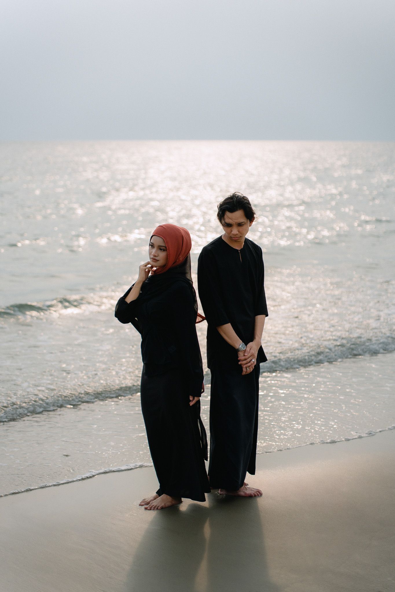 Couple in matching black outfits standing on a beach, woman wearing red hijab, ocean waves in background.