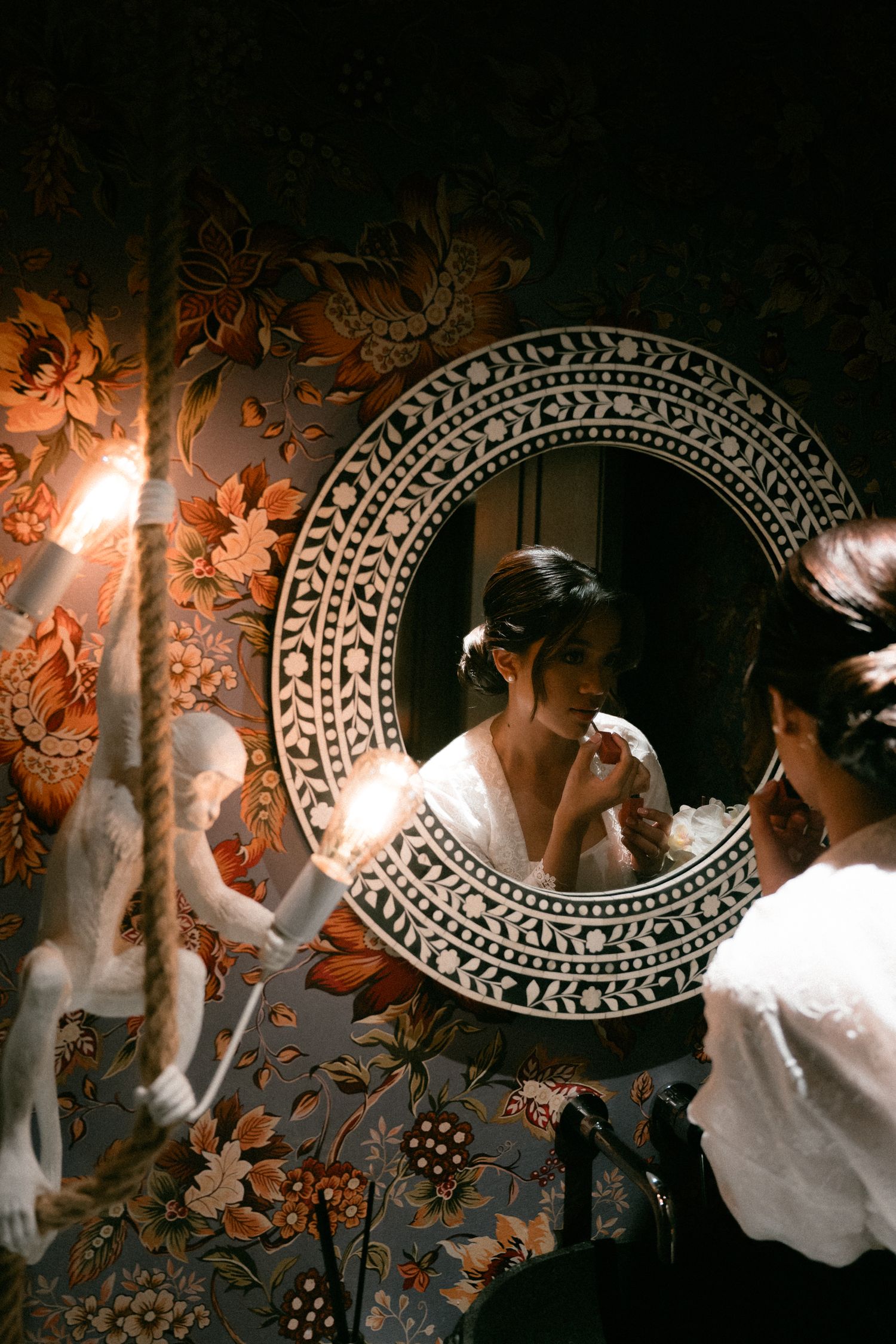 A vintage-style shot of an ornate round mirror against floral wallpaper with decorative cherub artwork and soft lighting.