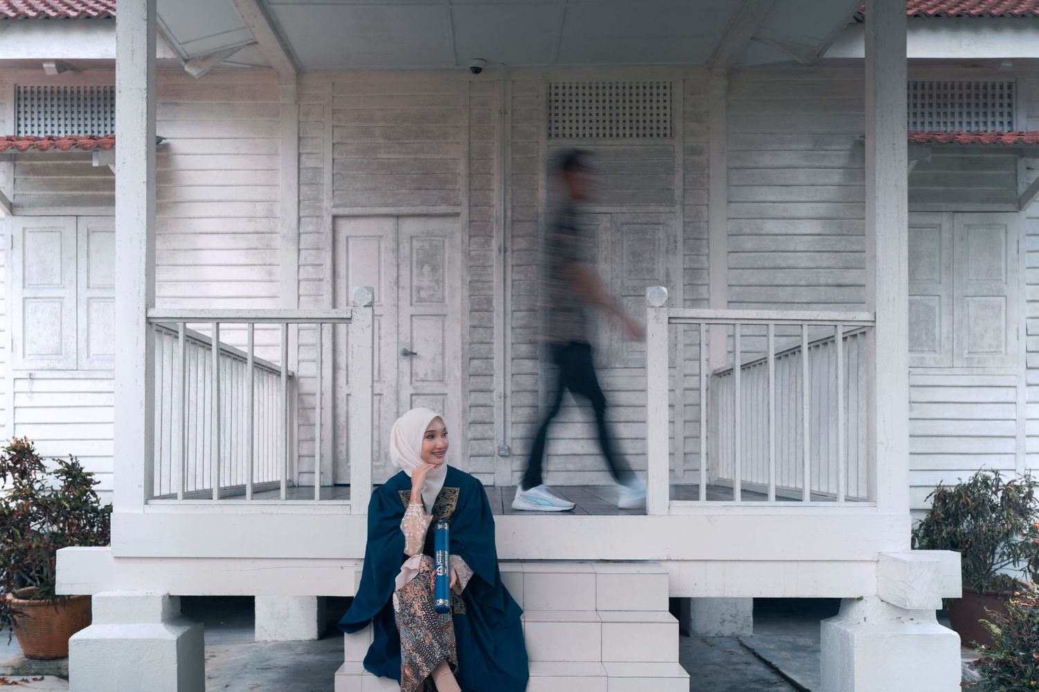 Graduate poses on white porch while blurred figure walks past.