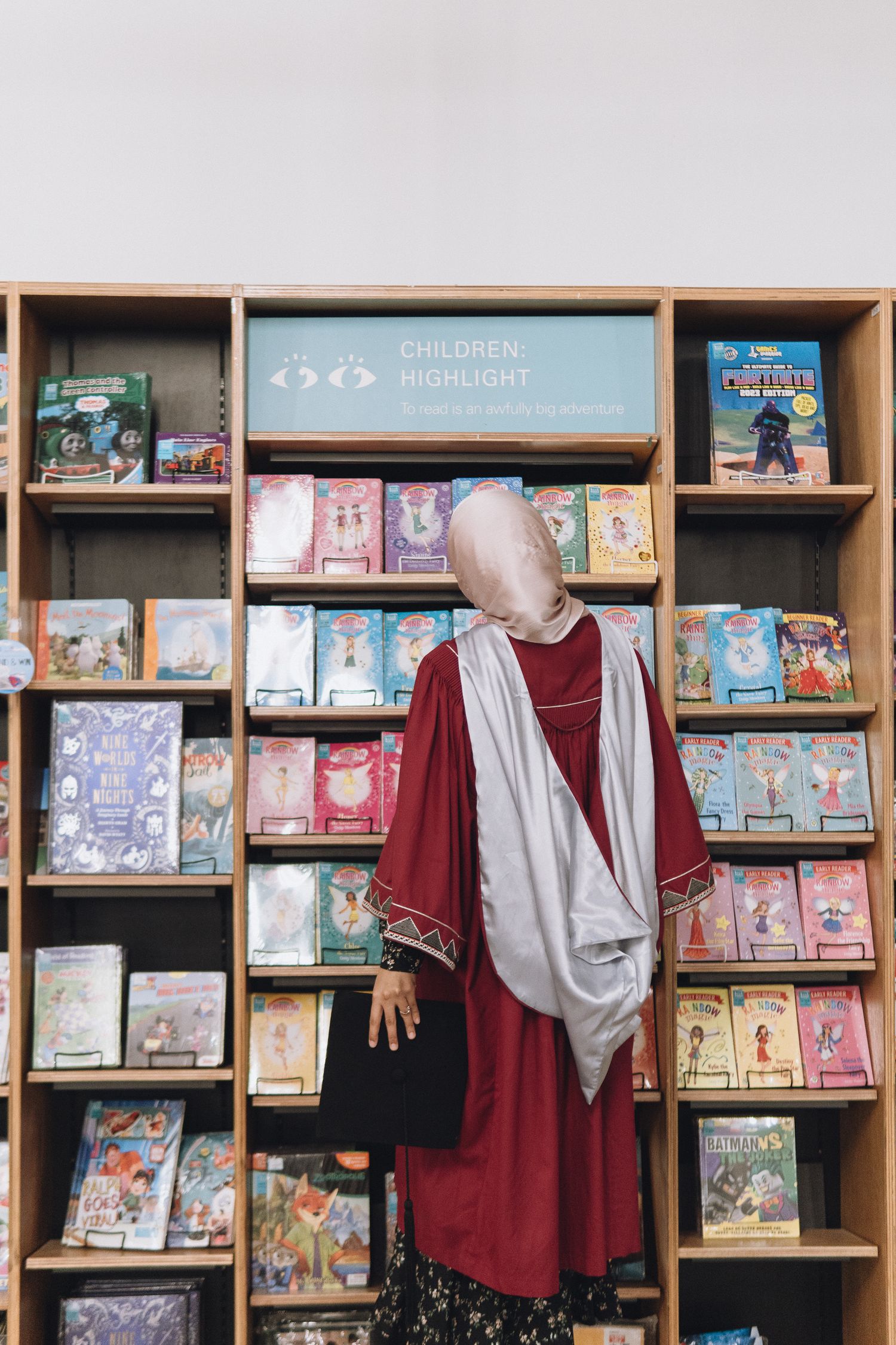 A graduate in red regalia examines children's books on wooden library shelves.