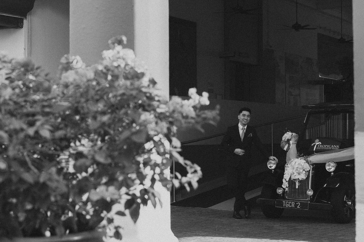 A vintage black and white photograph showing someone seated near a car entrance with flowering bushes in the foreground.