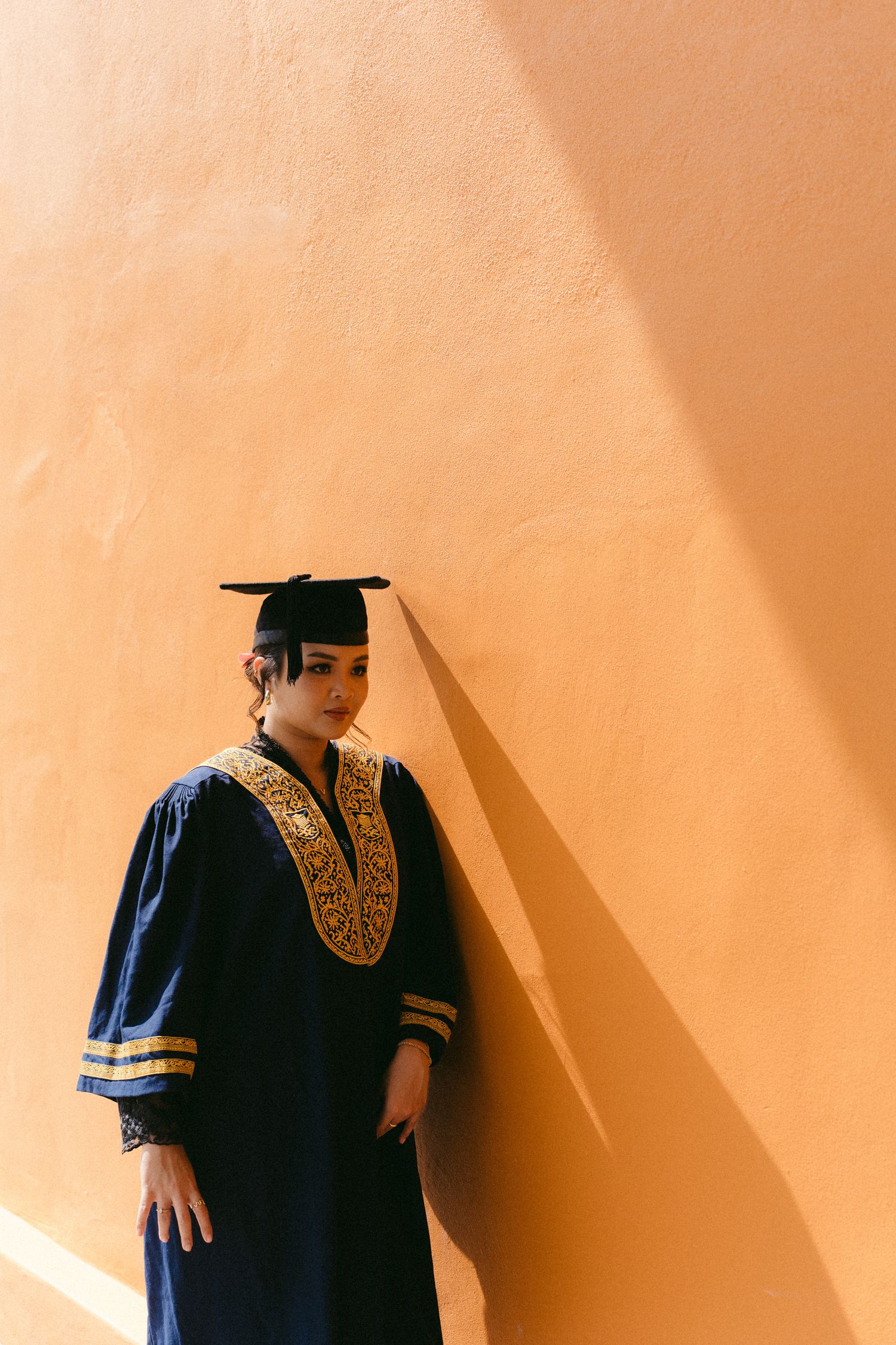 A graduate stands against a peach-colored wall wearing a navy blue gown with gold embroidery and a square academic cap.
