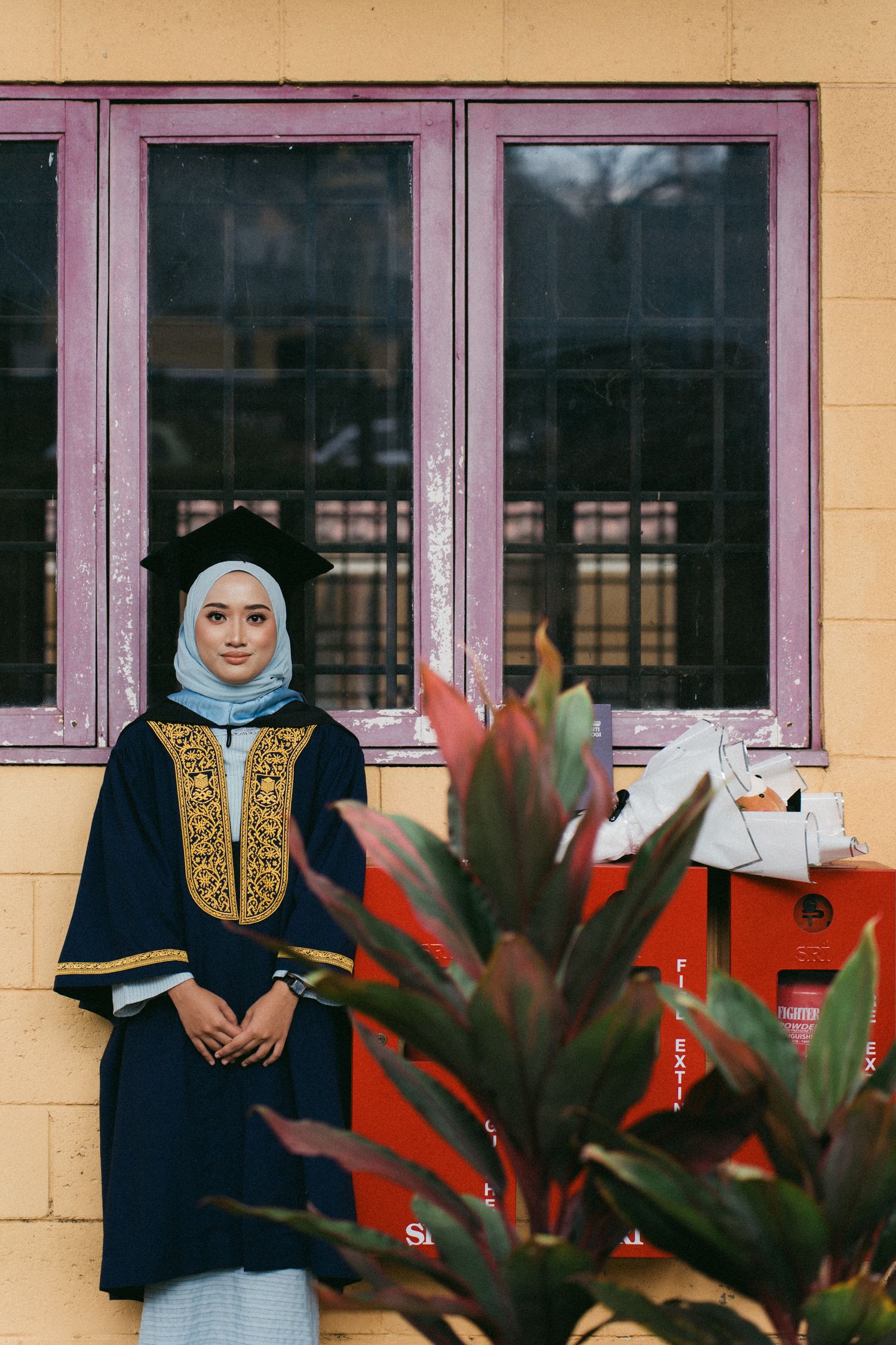 A person in a navy blue traditional dress with gold embroidery and light blue hijab stands against a yellow wall with windows.