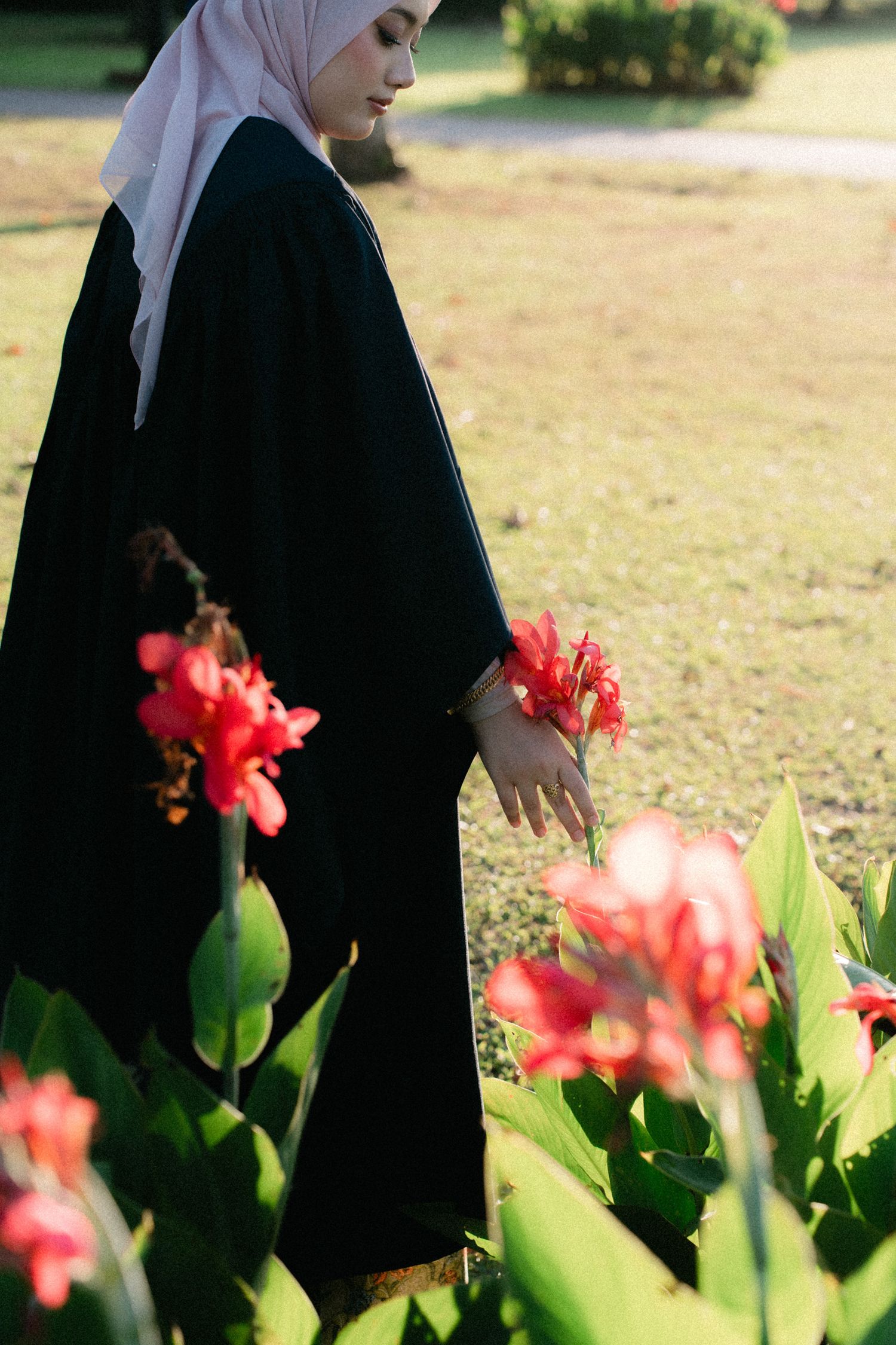 A person in black clothing and white hijab stands near vibrant red flowers and green foliage in a garden setting.