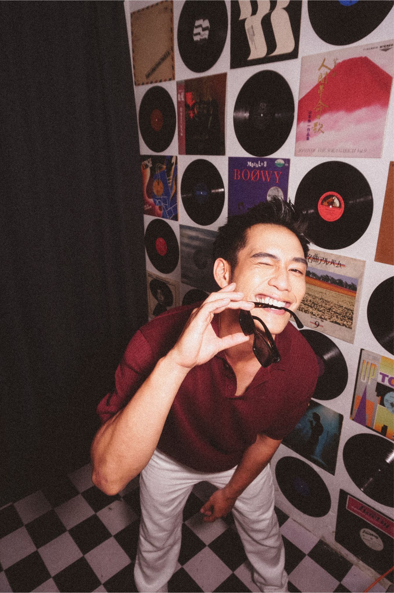 Smiling young man in burgundy sweater poses with headphones in front of vinyl record wall on checkered floor.