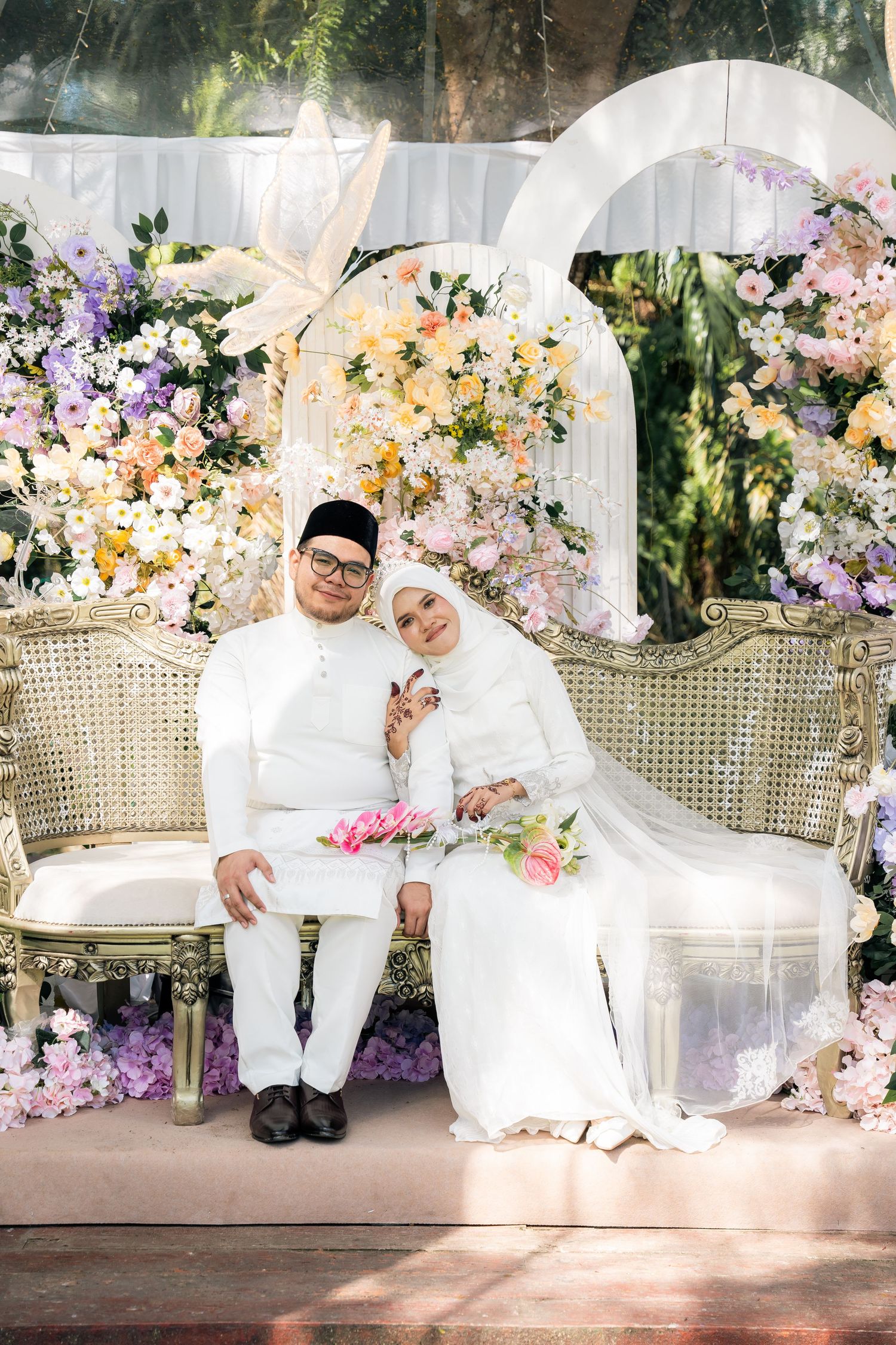 Beautiful wedding scene with couple seated on elegant chairs surrounded by pink, purple and yellow floral arrangements.
