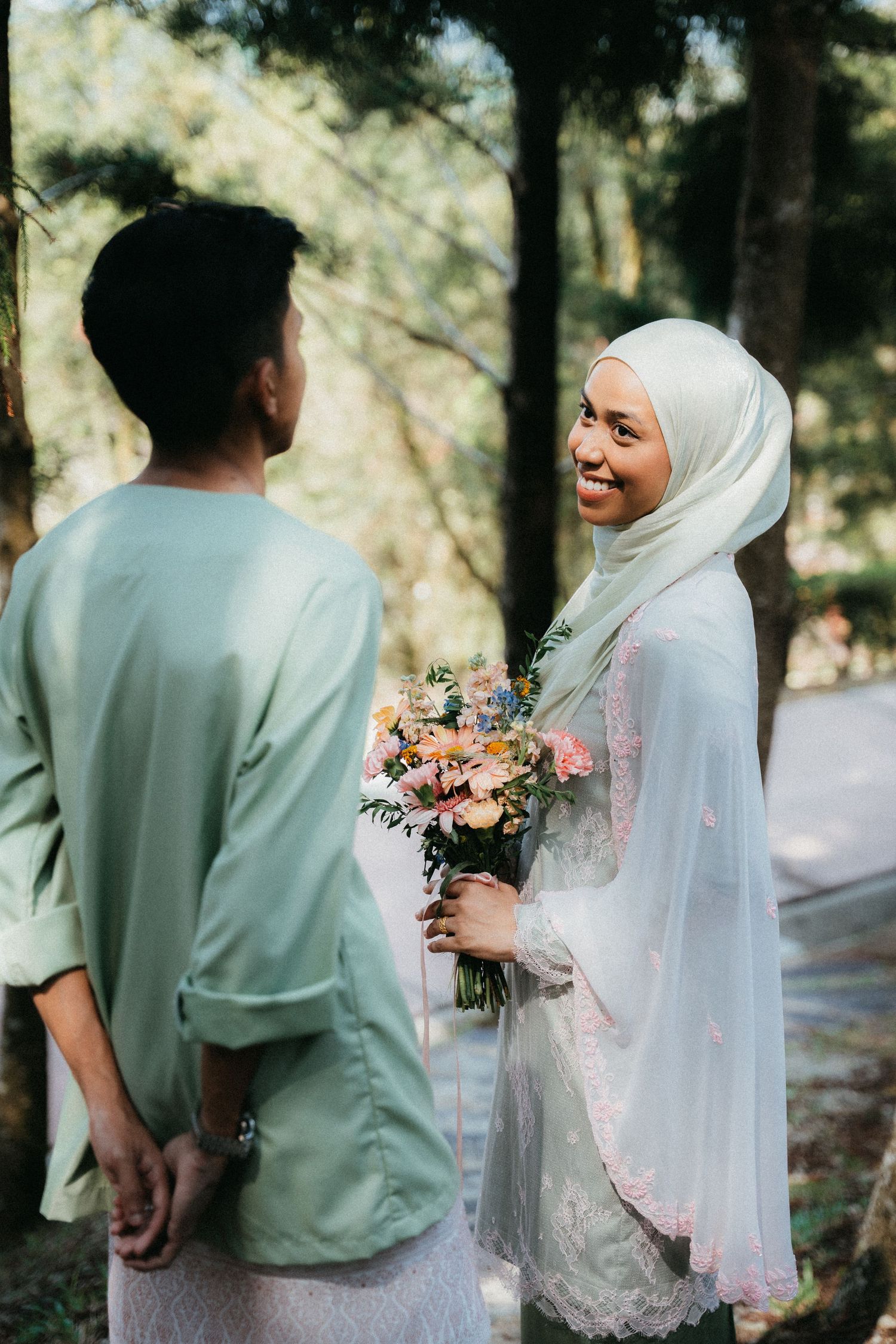 Romantic outdoor photoshoot showing couple in light green shirt and white hijab holding pink floral bouquet.