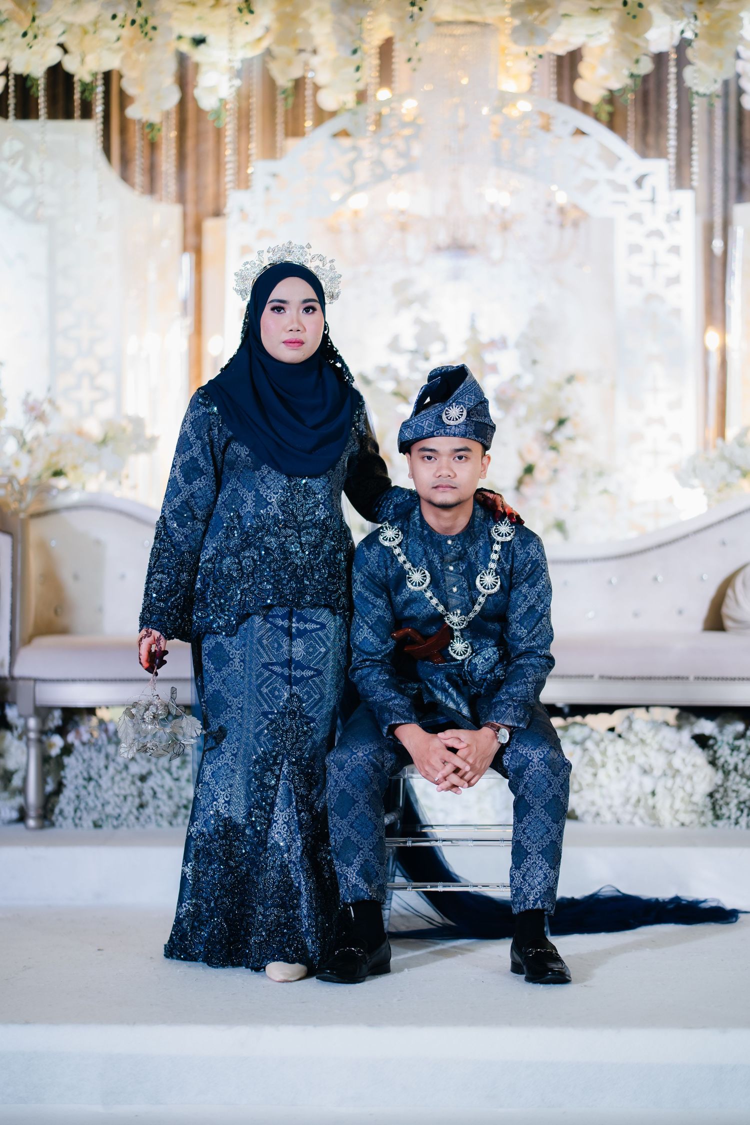 Two people wearing traditional blue Malay formal attire with matching patterns pose in an elegantly decorated setting.
