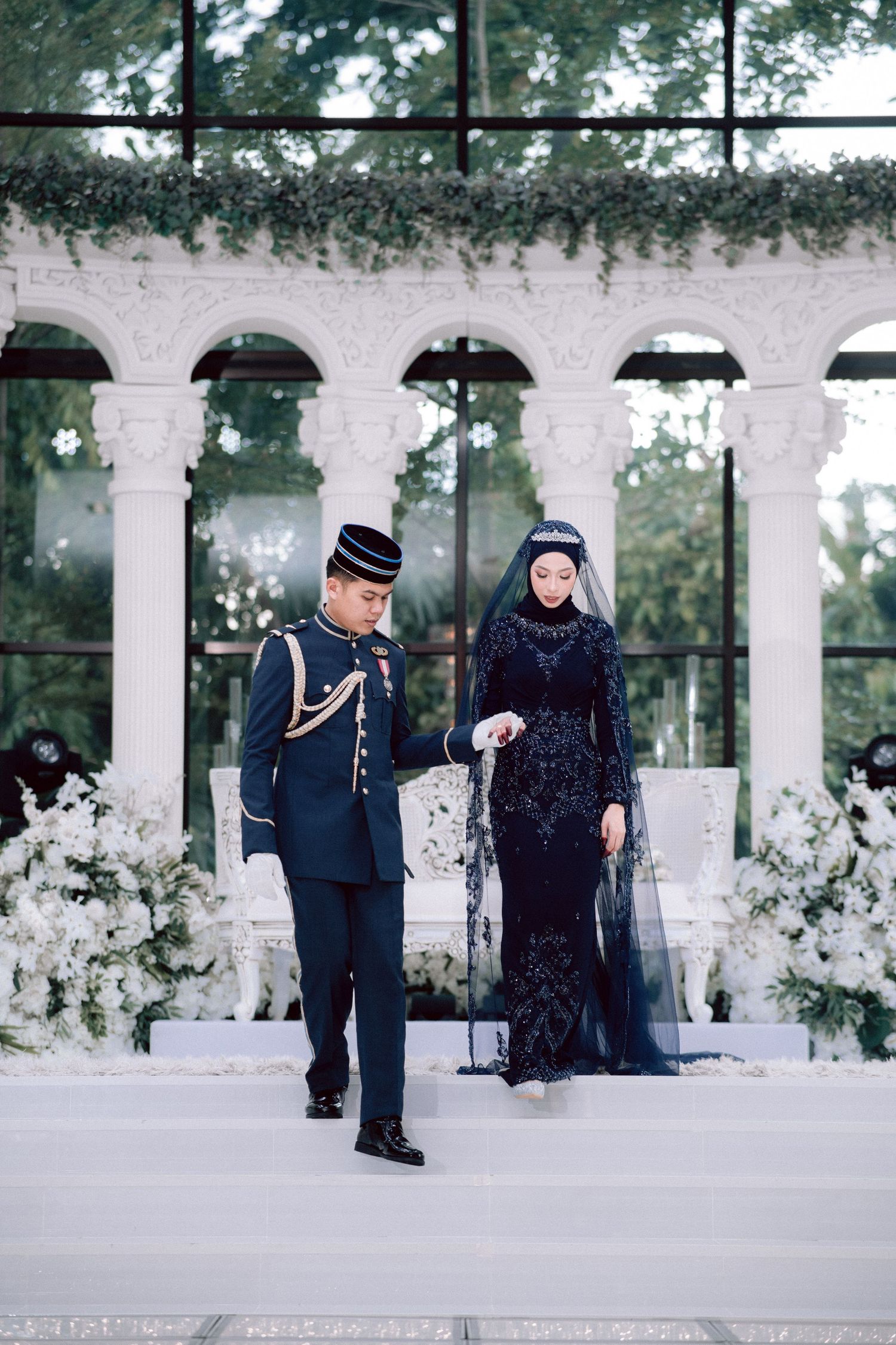 Military officer in formal uniform walks beside elegantly dressed figure in dark blue gown under white columned architecture.