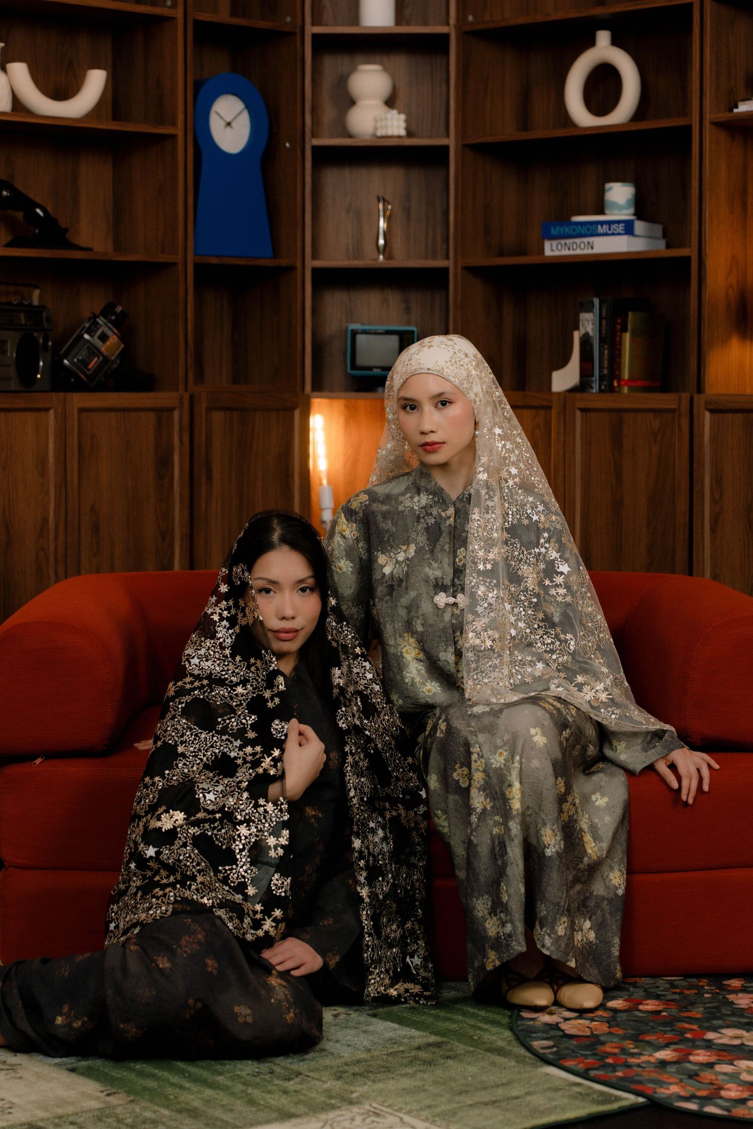Two people wearing ornate veils sit on a red couch in front of wooden shelving with decorative objects.