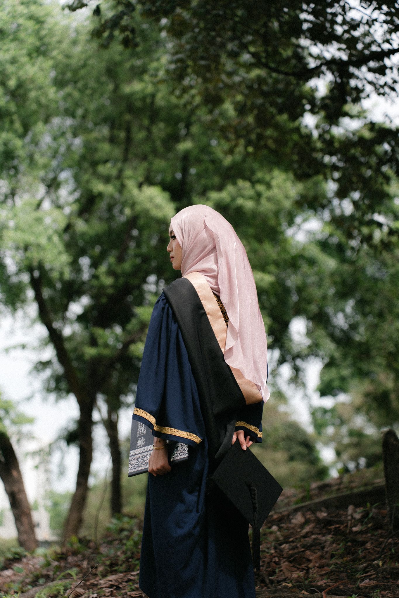 Muslim woman in pink hijab and navy graduation gown holding mortarboard outdoors among green trees.