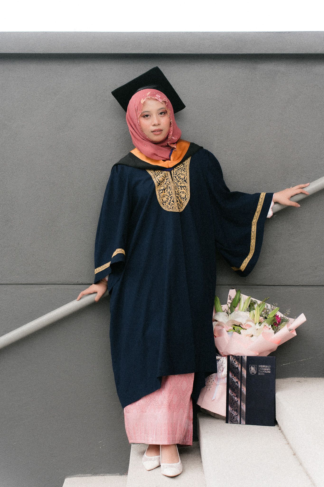 Muslim woman in graduation gown and pink hijab holding flower bouquet, celebrating academic achievement.