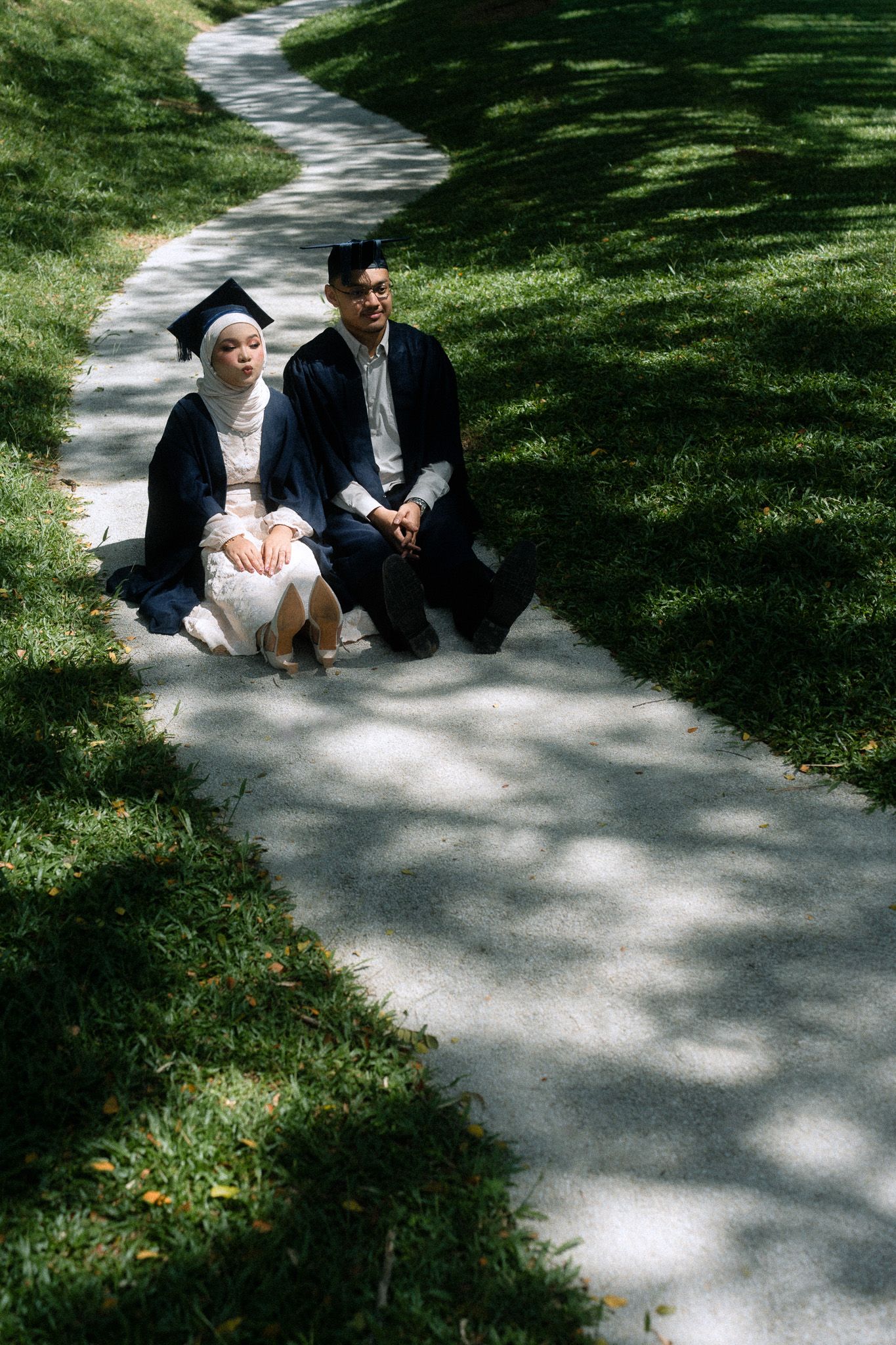 Two graduates in caps and gowns sit together on a winding campus pathway surrounded by lush green grass.