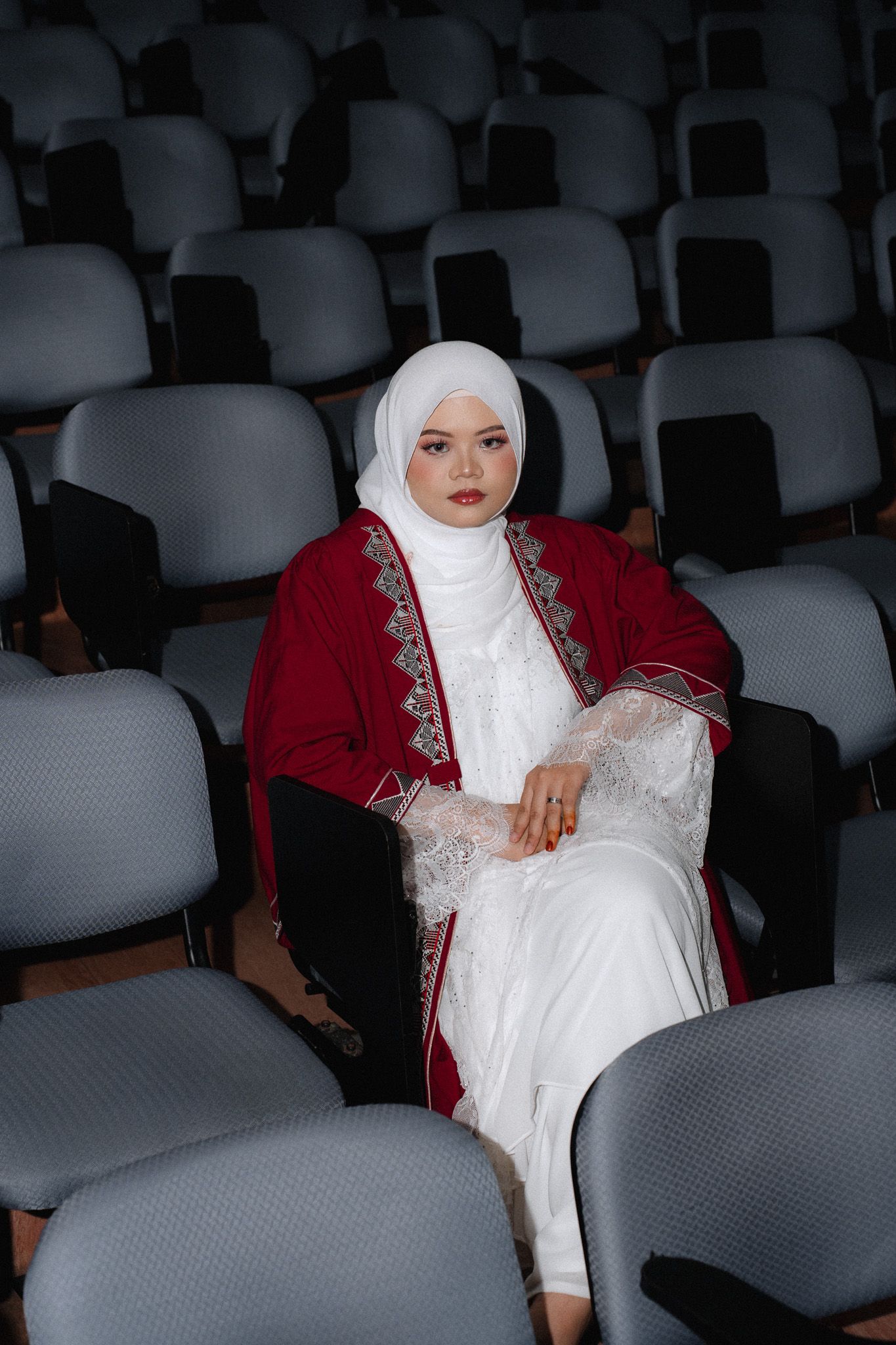 Woman in white hijab and red embroidered jacket sits alone in rows of gray auditorium seats.