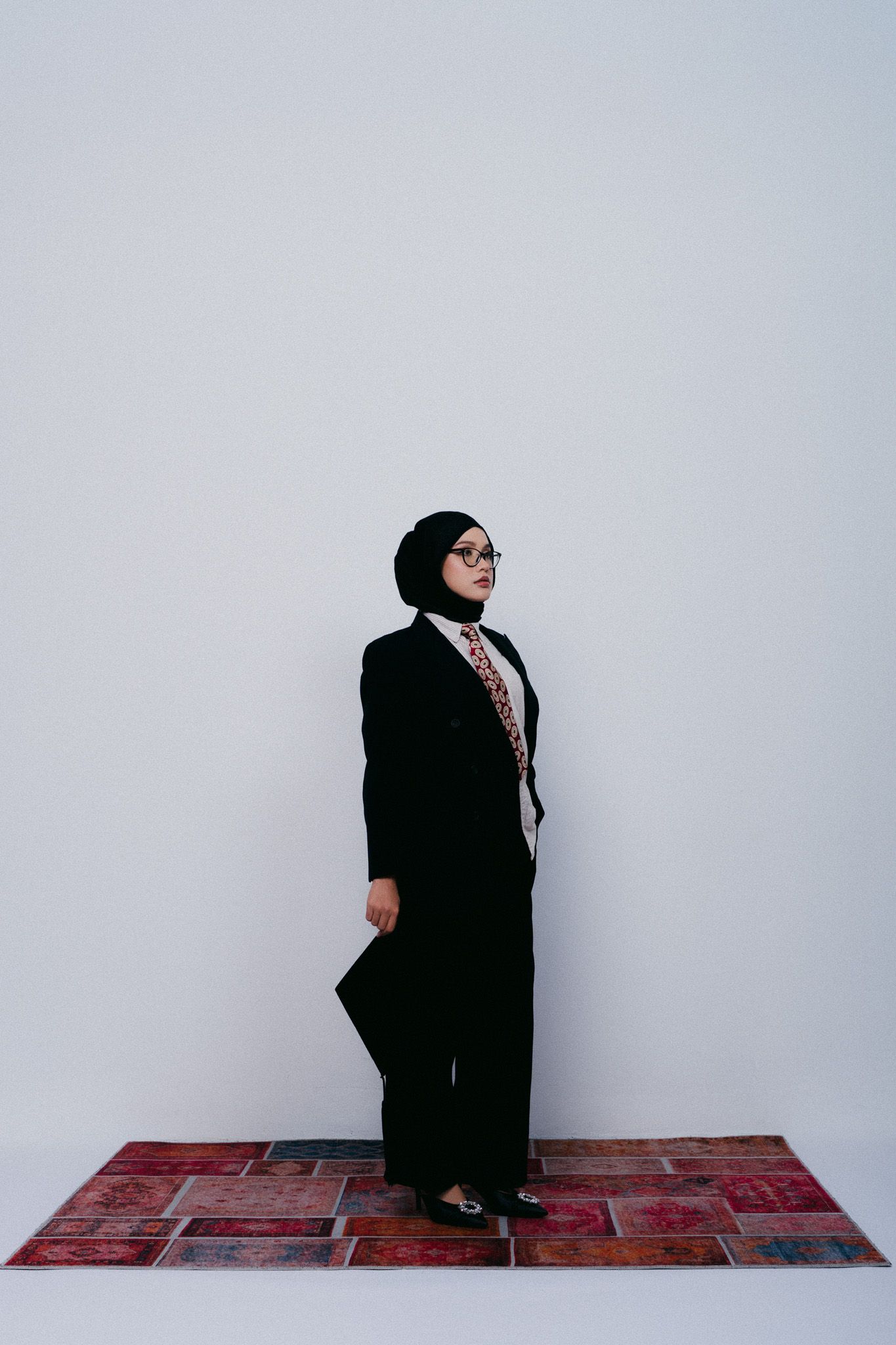 A professional woman in a black hijab, blazer, and wide-leg pants stands on a patterned rug holding a folder.